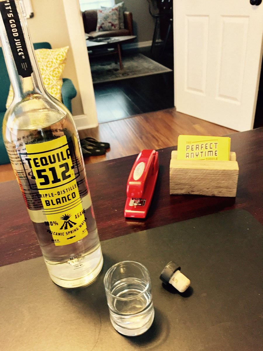 drinks 桌布 壁紙 — 一個透明的龍舌蘭白瓶放在一張深色桌子上，旁邊有一個杯子和一個紅色釘書機連木製底座。