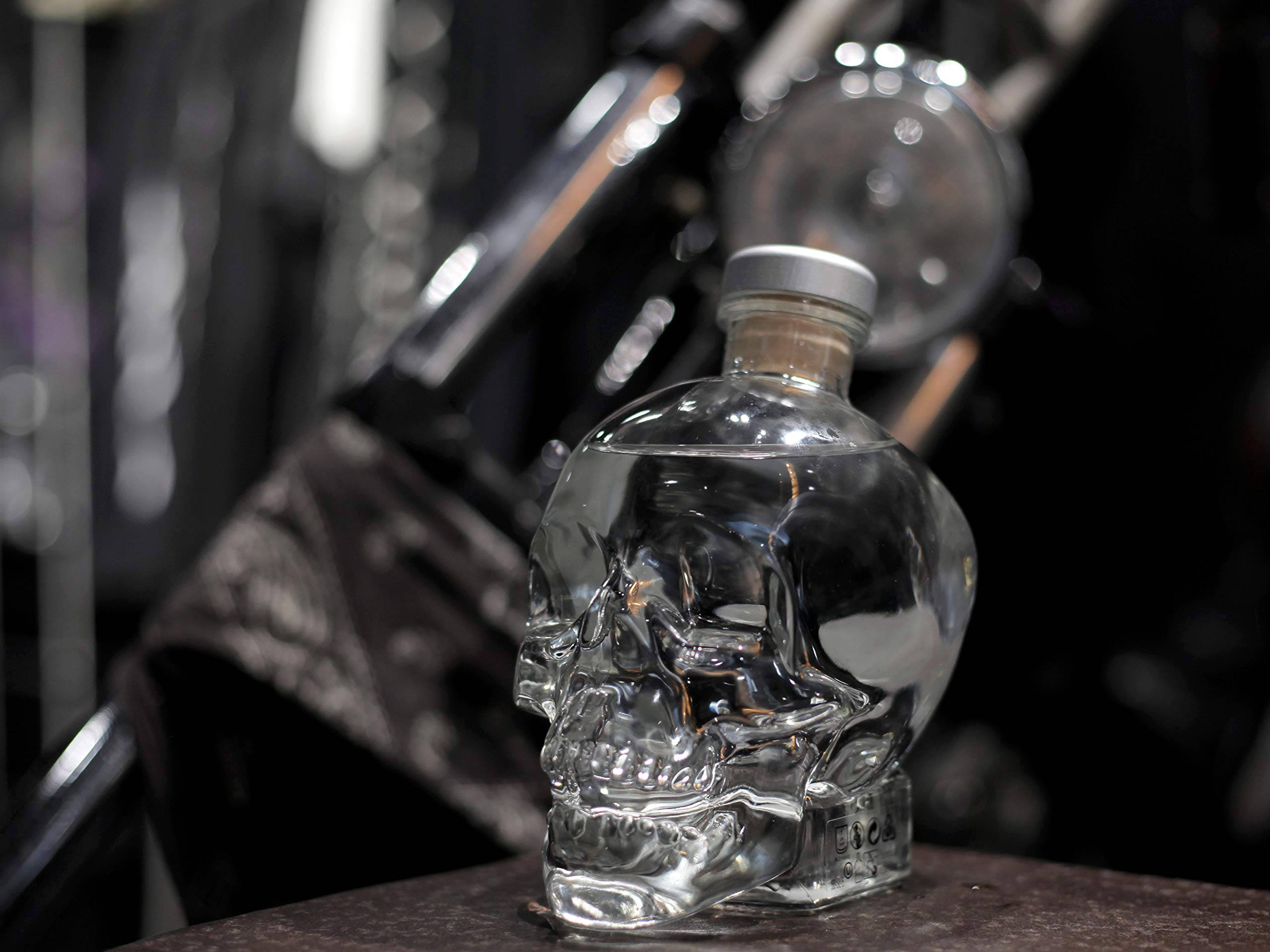 Drink 桌布 壁紙 — 一個透明骷髏形狀的伏特加酒瓶放在黑暗的表面上，背景有模糊的機車零件。