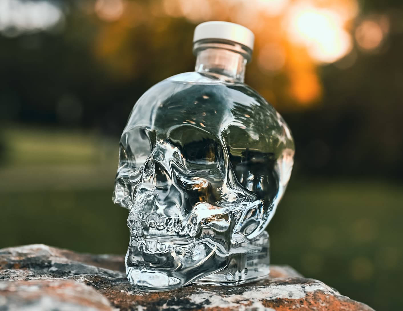 Drink 桌布 壁紙 — 一個透明骷髏形狀的伏特加酒瓶放在一塊長滿苔蘚的石頭上，背景是溫暖的金色散景。