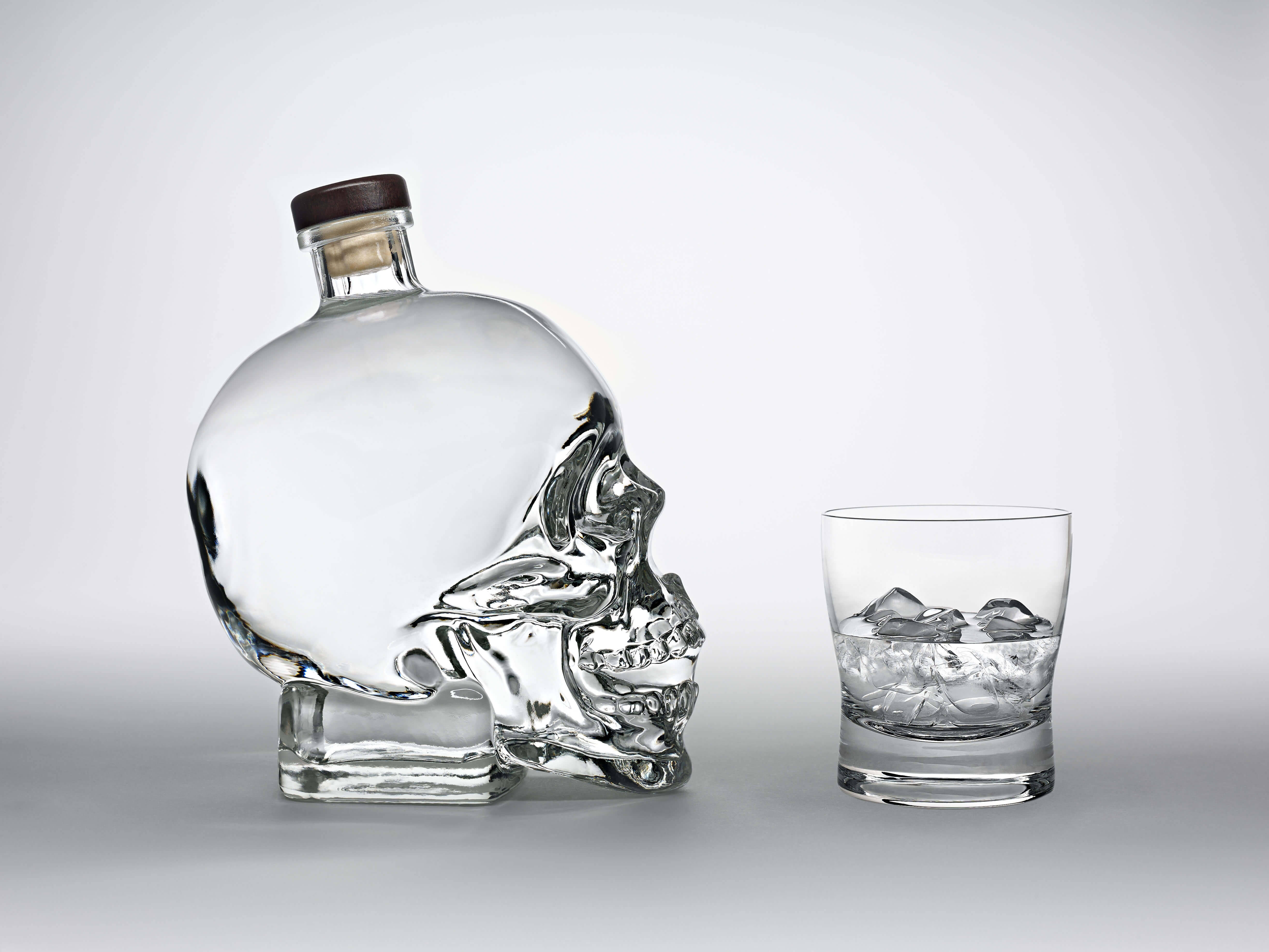 Drink 桌布 壁紙 — 一個透明骷髏形狀的酒瓶，帶有木蓋，旁邊放著一個裝滿冰塊的岩石杯，背景為中性灰色。
