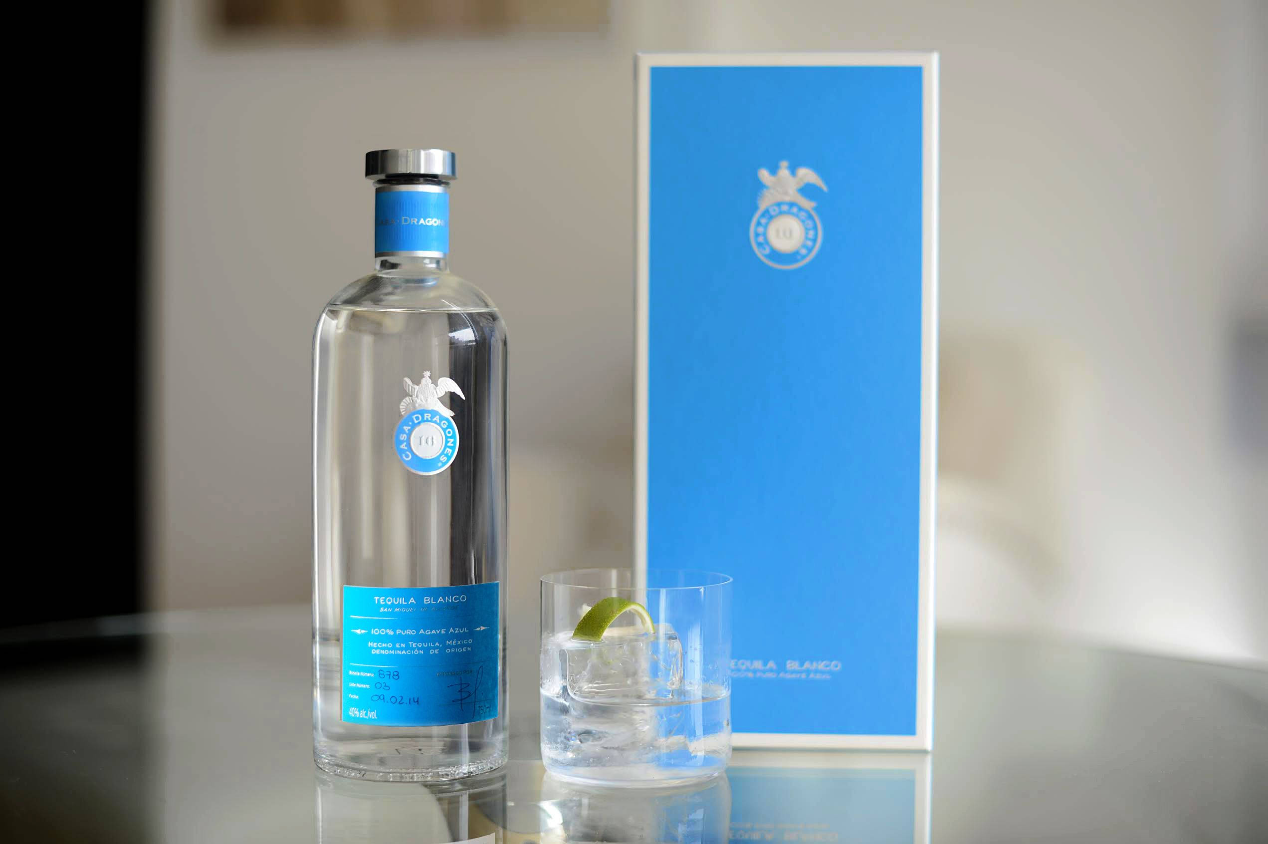 cocktail 桌布 壁紙 — 一個透明龍舌蘭酒瓶，帶有藍色標籤和盒子，放在反射桌面上，旁邊有一個加冰和萊姆角的玻璃杯。
