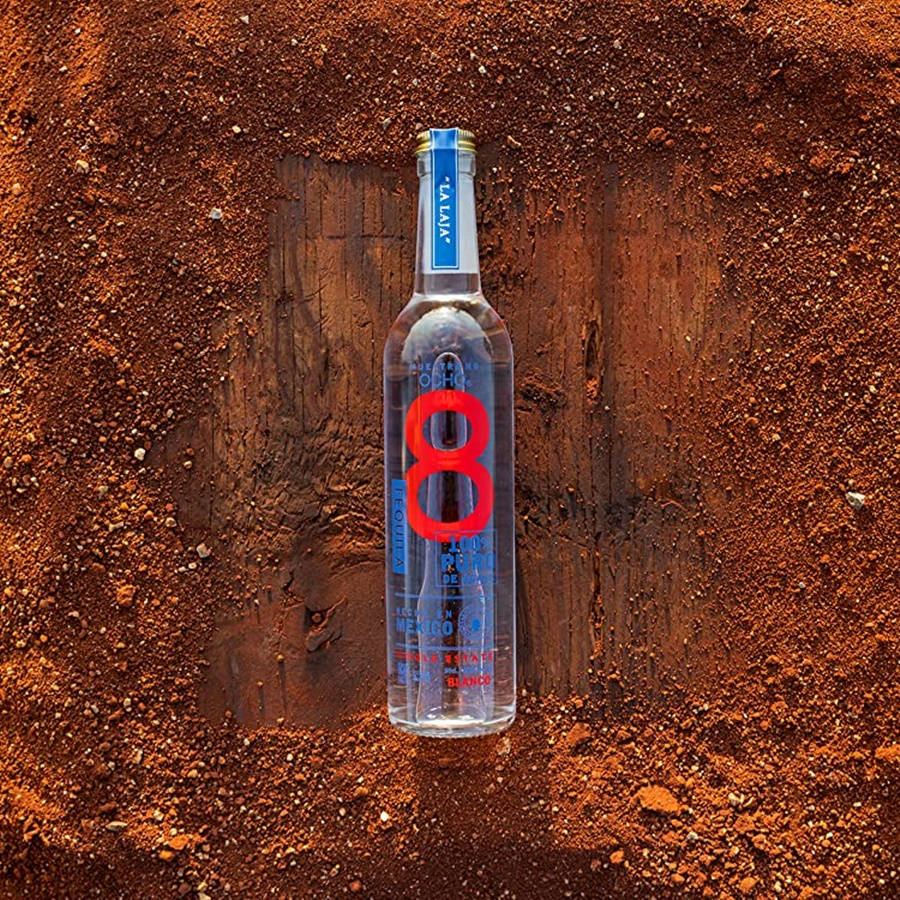 bottle 桌布 壁紙 — 一個透明龍舌蘭酒瓶，帶有醒目的紅色數字八標籤，直立於豐富的紅棕色土壤中，背景為風化的木板。
