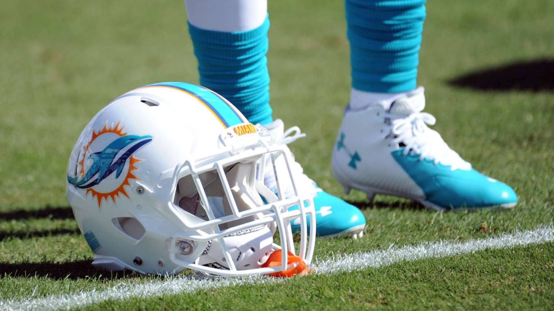 dolphins 桌布 壁紙 — 一個邁阿密海豚隊美式足球頭盔放在綠色草地上，旁邊是球員的青綠色和白色釘鞋。