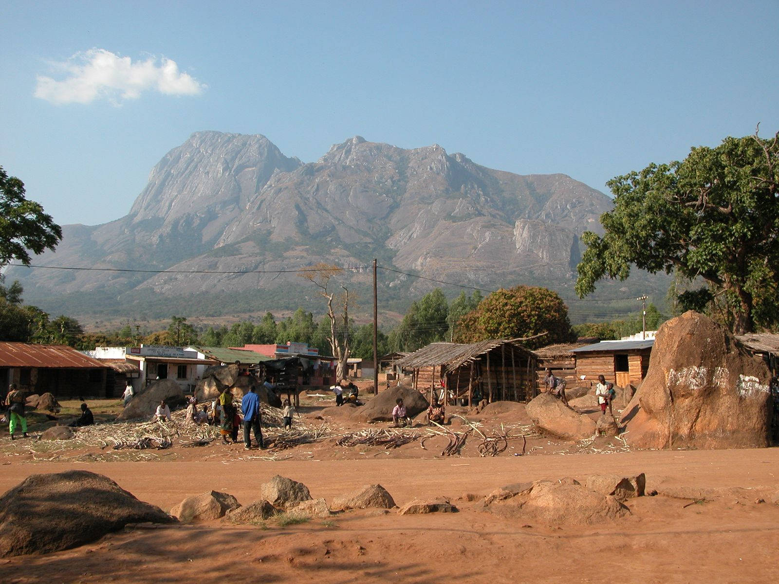 Malawi 桌布 壁紙 — 一個鄉村村莊，有泥屋和人們在廣闊的藍天下工作，背景中有一座巨大的岩石山聳立。