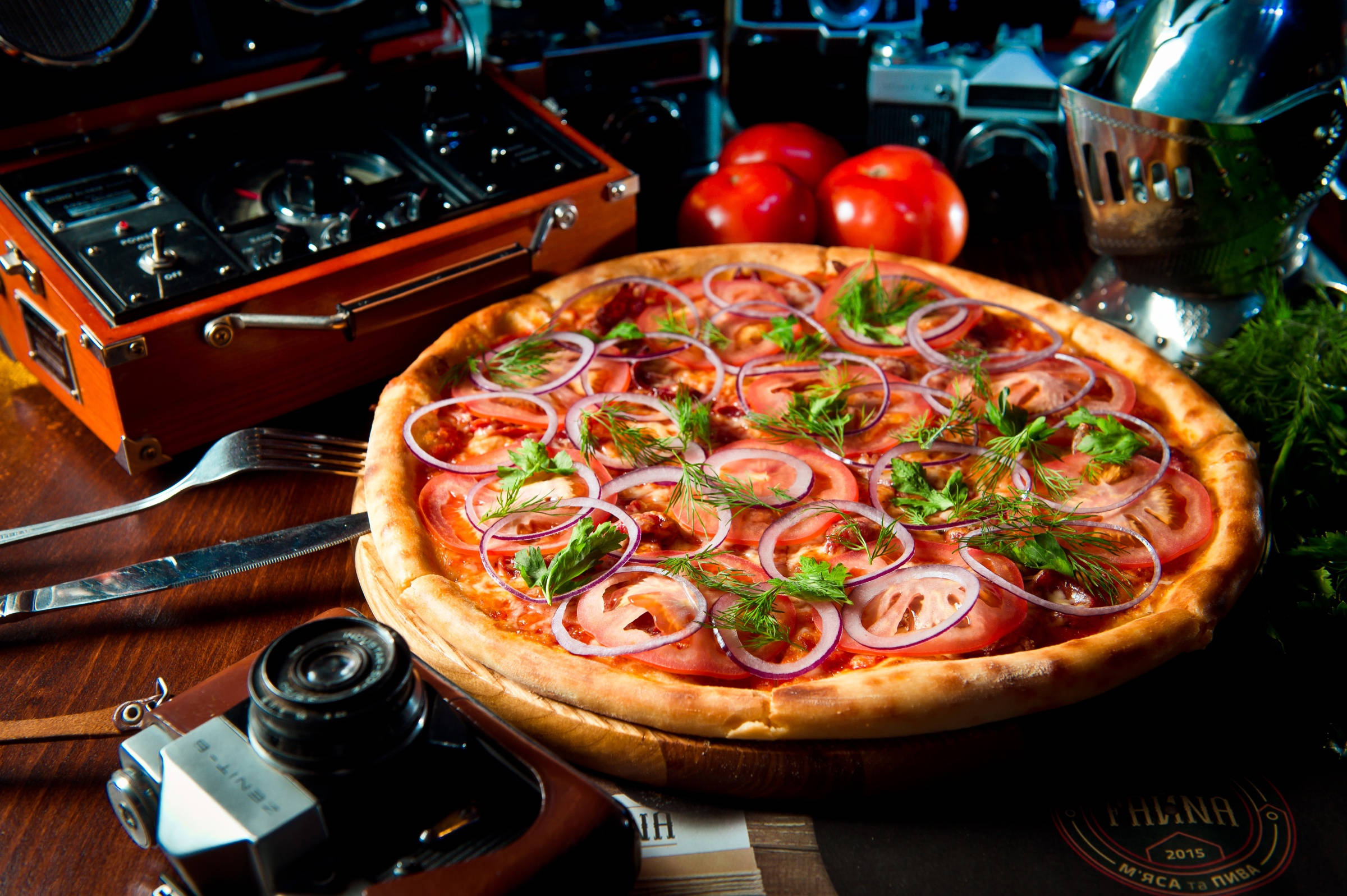 Pizza 桌布 壁紙 — 一個鄉村風格的披薩，上面鋪有番茄片、紅洋蔥和新鮮香草，放在木桌上，周圍環繞著復古相機和番茄。