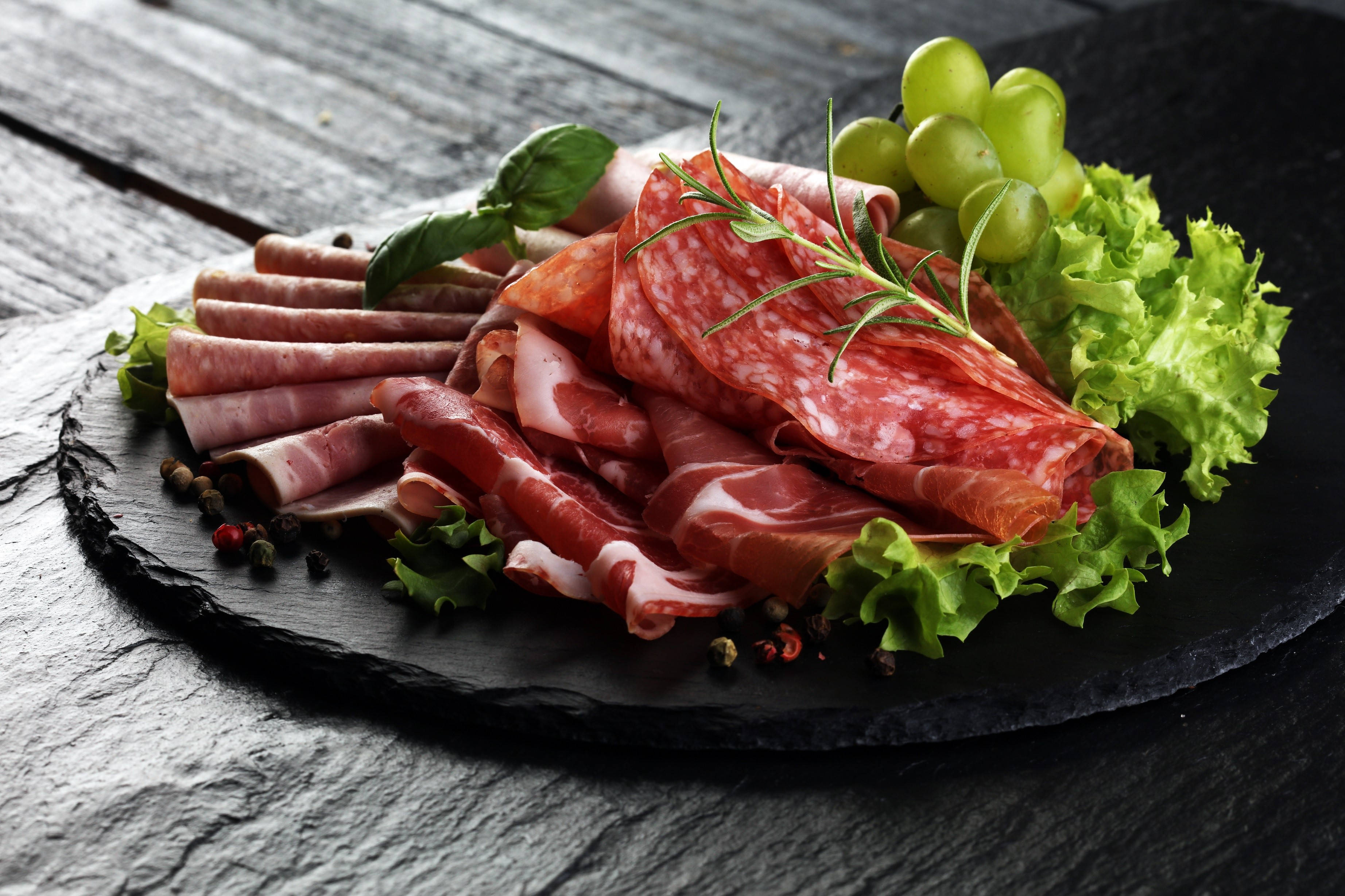 meat 桌布 壁紙 — 一個鄉村風格的熟食拼盤，上面擺放著醃製肉類、葡萄、生菜和香草，放在深色石板板上。