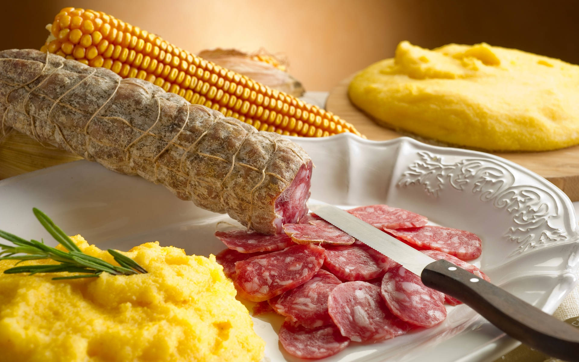 Polenta 桌布 壁紙 — 一個鄉村風格的盤子，上面放著切片薩拉米、整粒玉米和奶油玉米粥，並用迷迭香點綴，在溫暖的燈光下。
