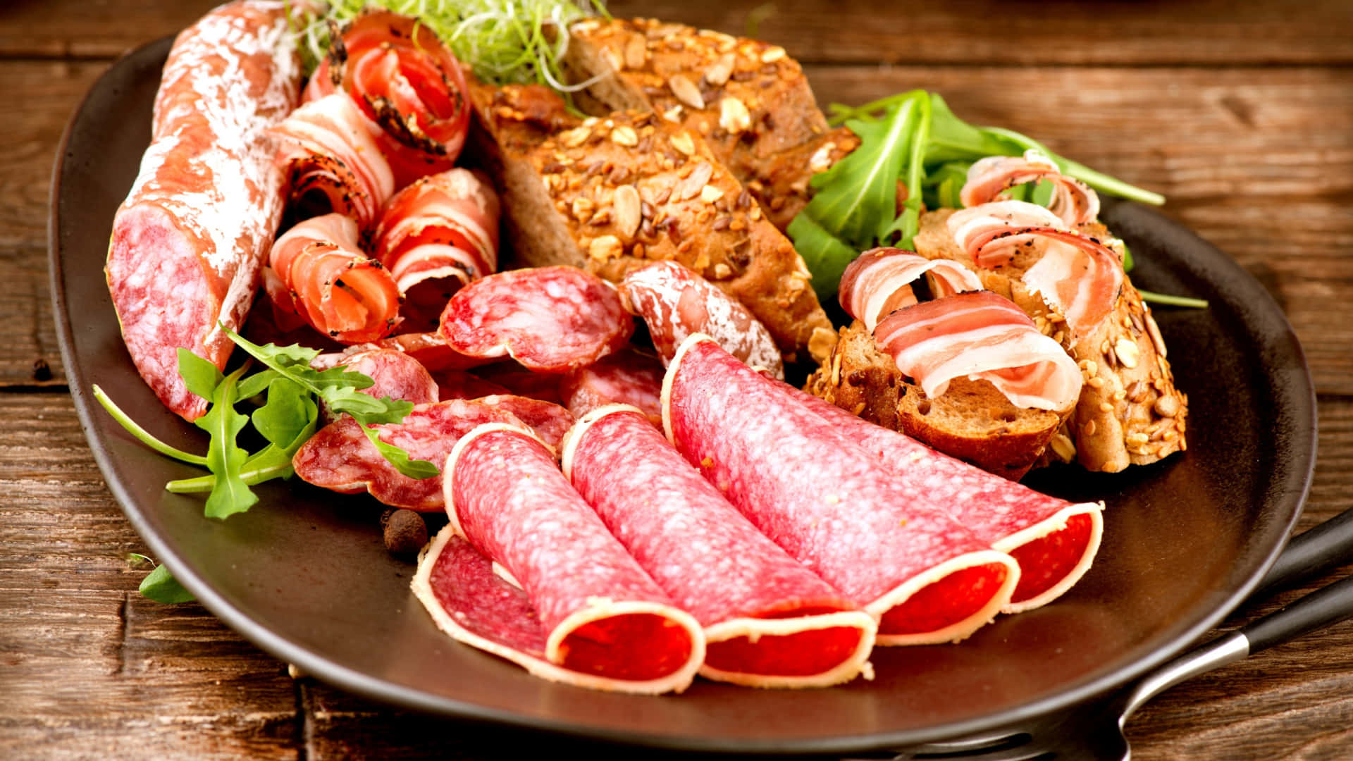 Red Meat 桌布 壁紙 — 一個鄉村風格的盤子，堆滿了醃肉、芝麻菜和種子麵包，放在一張風化的木桌上。