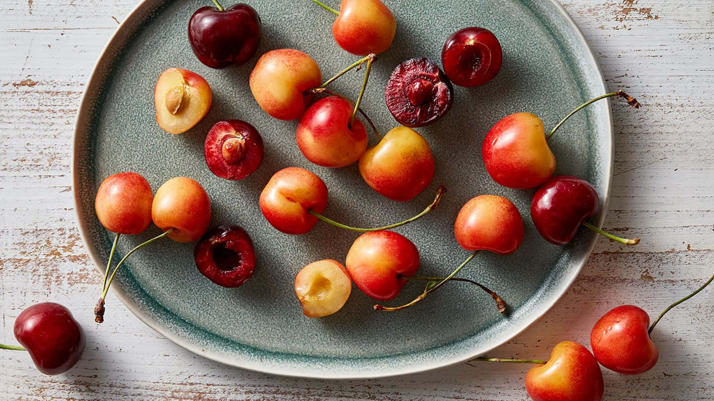 Red Cherries 桌布 壁紙 — 一個鄉村風格的盤子上放著色彩鮮豔的完整和半切櫻桃，置於經風化的白色木質表面上。
