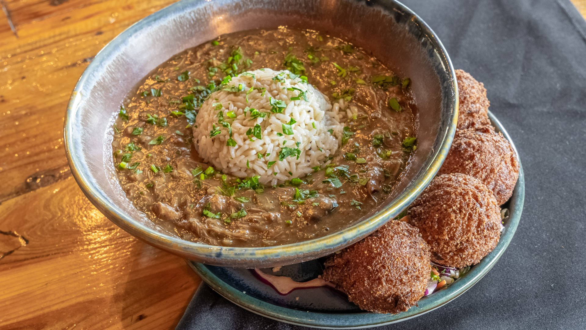 meatball 桌布 壁紙 — 一個鄉村風格的碗，盛滿濃郁的棕色燉菜，上面放著一堆白米和新鮮香草，旁邊是金黃色的炸餃子，放在木桌上。