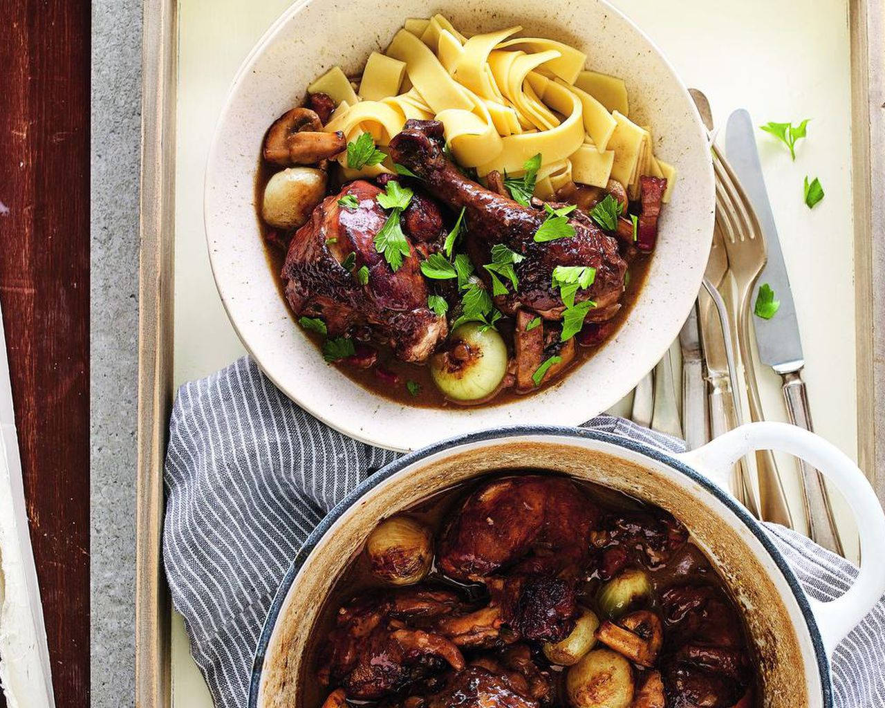 Coq Au Vin 桌布 壁紙 — 一個鄉村風格的碗，盛載著豐富的鴨腿油封和寬麵條及新鮮歐芹，旁邊有一個鑄鐵鍋放在條紋布上。