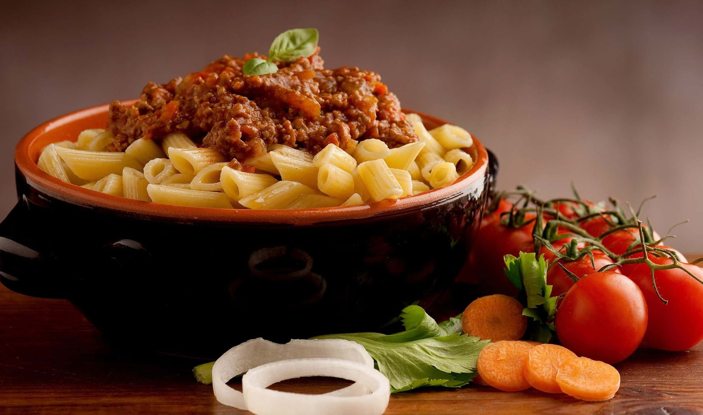 2440x1440 Pasta 桌布 壁紙 — 一個鄉村風格的碗，盛載著長通粉，上面鋪滿濃郁的肉醬和新鮮羅勒，旁邊放著鮮艷的櫻桃番茄和切片蔬菜，置於木桌上。