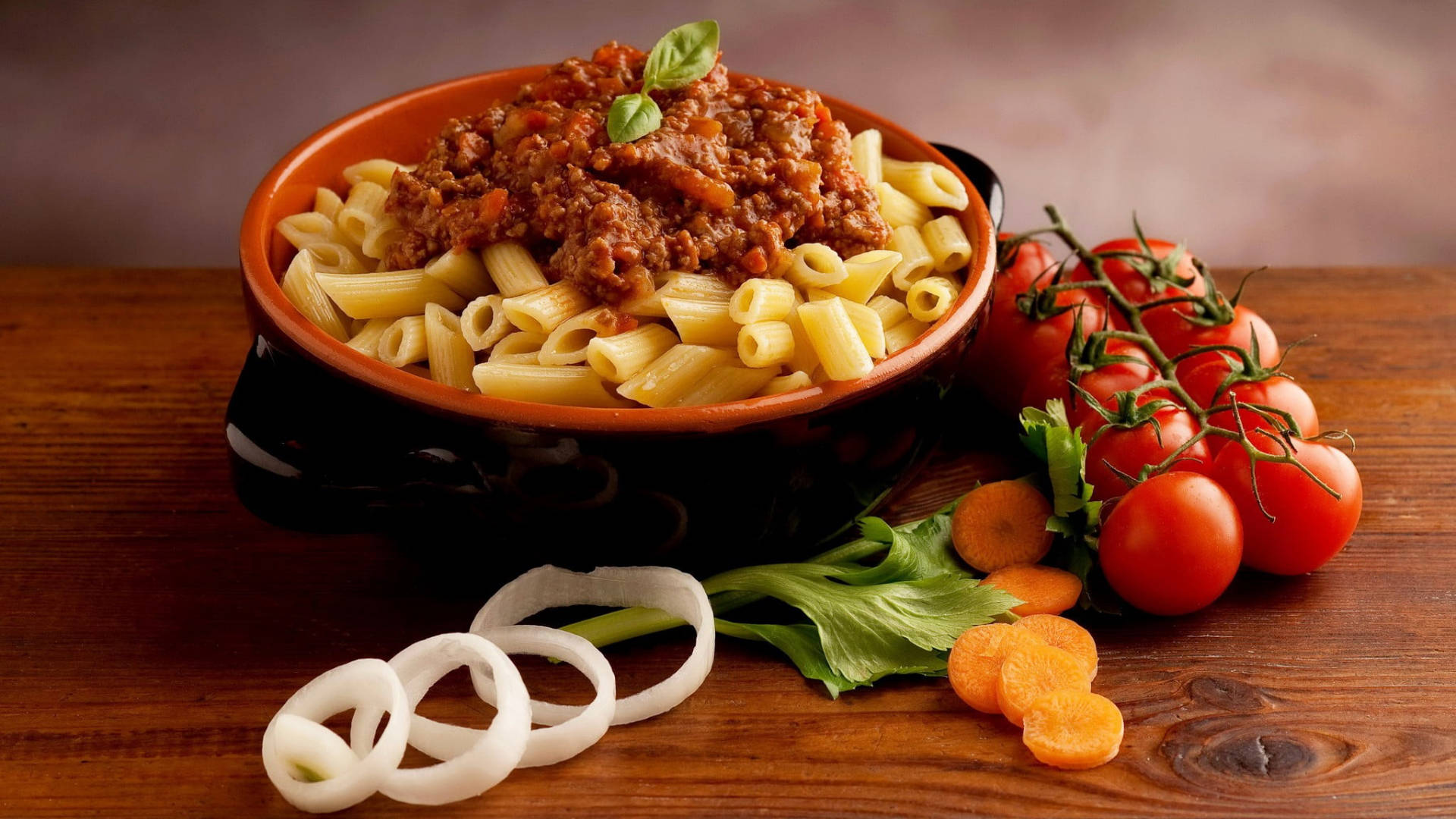 Pasta 桌布 壁紙 — 一個鄉村風格的碗，裡面裝著筆管麵，上面鋪滿濃郁的肉醬和新鮮羅勒，旁邊放著鮮豔的櫻桃番茄和切片洋蔥，在木桌上。
