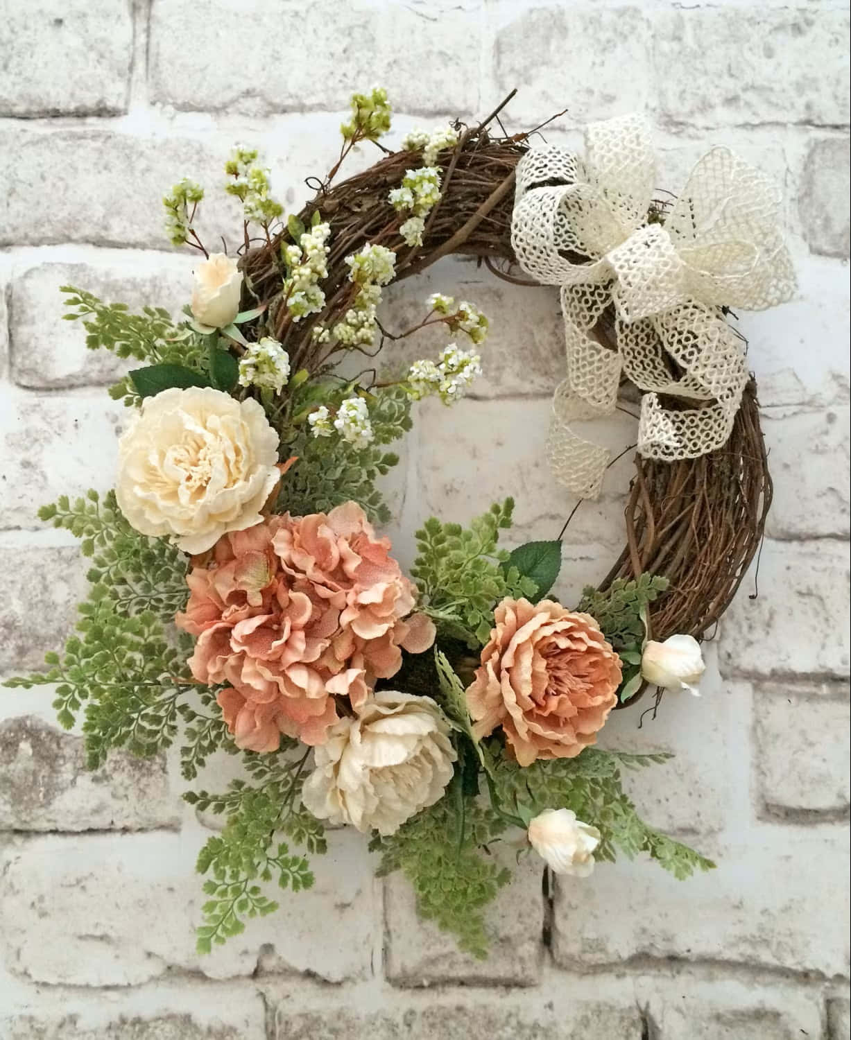 Spring Wreaths 桌布 壁紙 — 一個鄉村風格的花環，飾有蜜桃奶油玫瑰和綠色蕨類植物，搭配蕾絲蝴蝶結，掛在白色磚牆上。