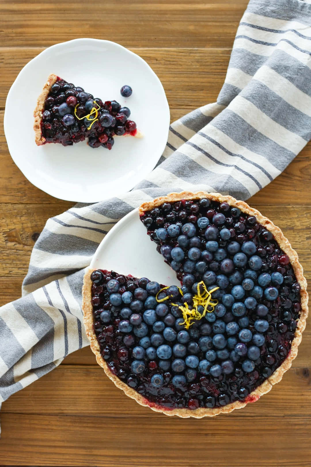 Blueberries Tart 桌布 壁紙 — 一個鄉村風格的藍莓塔，切片放在白色盤子上，旁邊有條紋布，置於木桌上。
