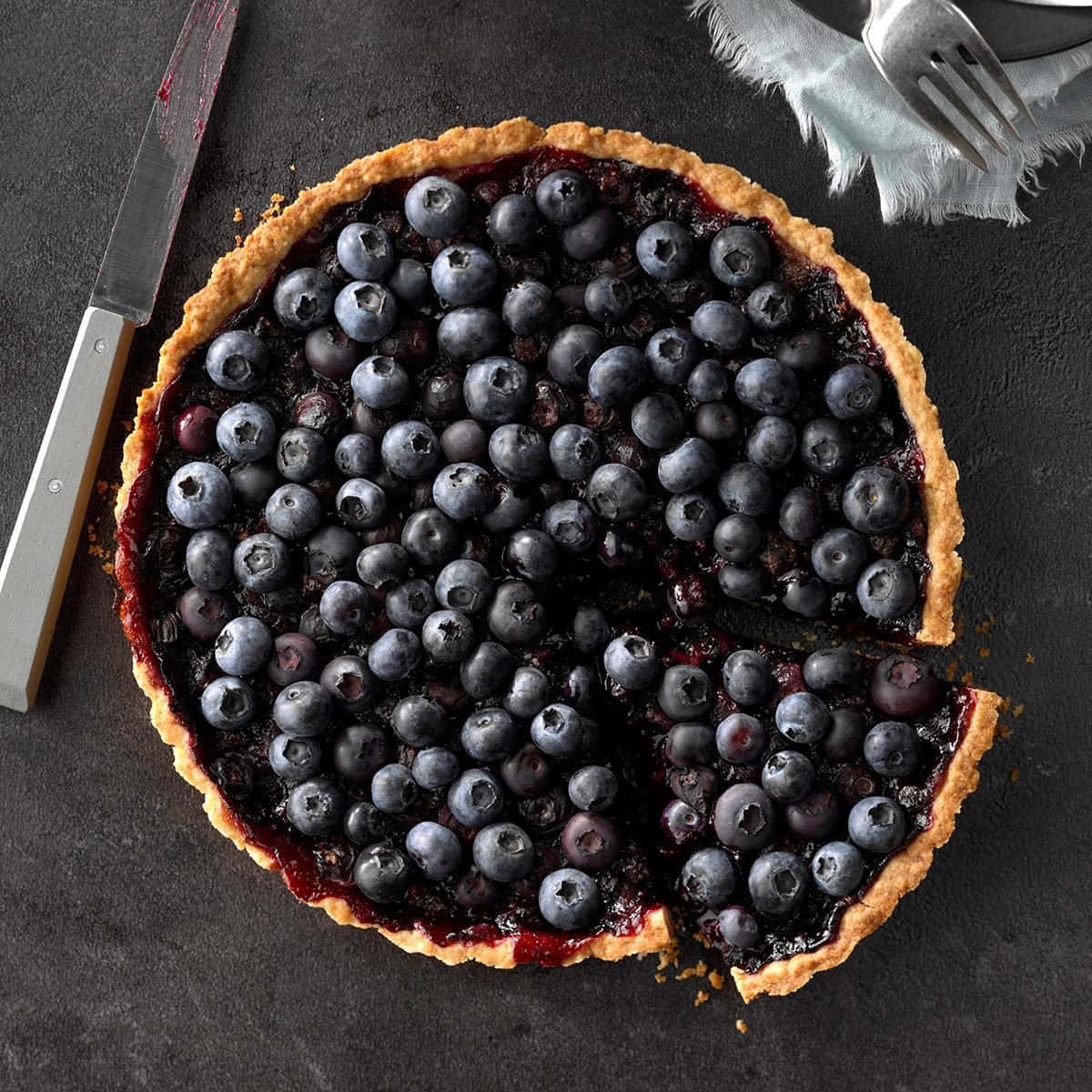 Blueberries Tart 桌布 壁紙 — 一個鄉村風格的藍莓塔，金黃色的外皮，放在深色石板表面上，附近有刀和叉。