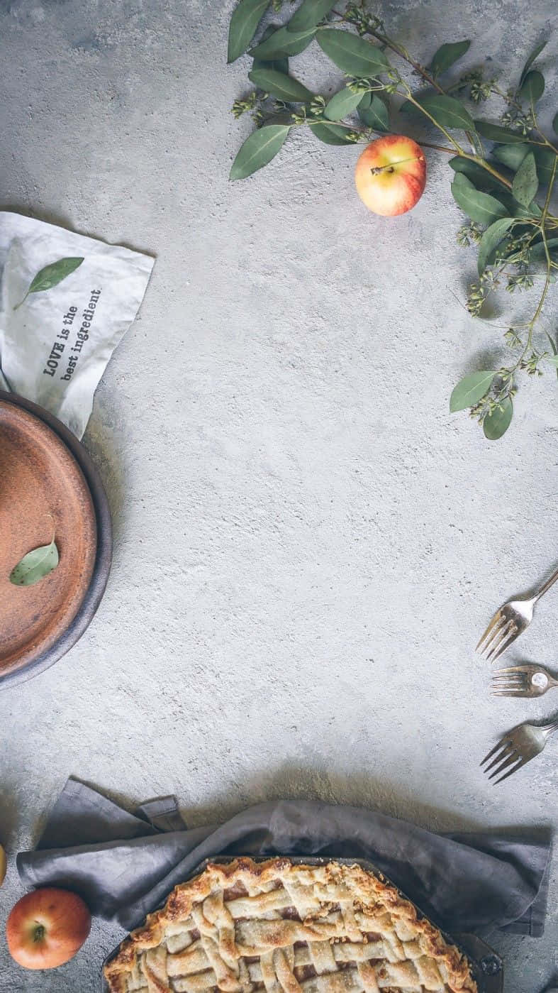 Android Kitchen 桌布 壁紙 — 一個鄉村風格的蘋果派，帶有格子狀外皮，放在深色布上，旁邊是新鮮蘋果和綠色植物，置於有紋理的灰色表面上。