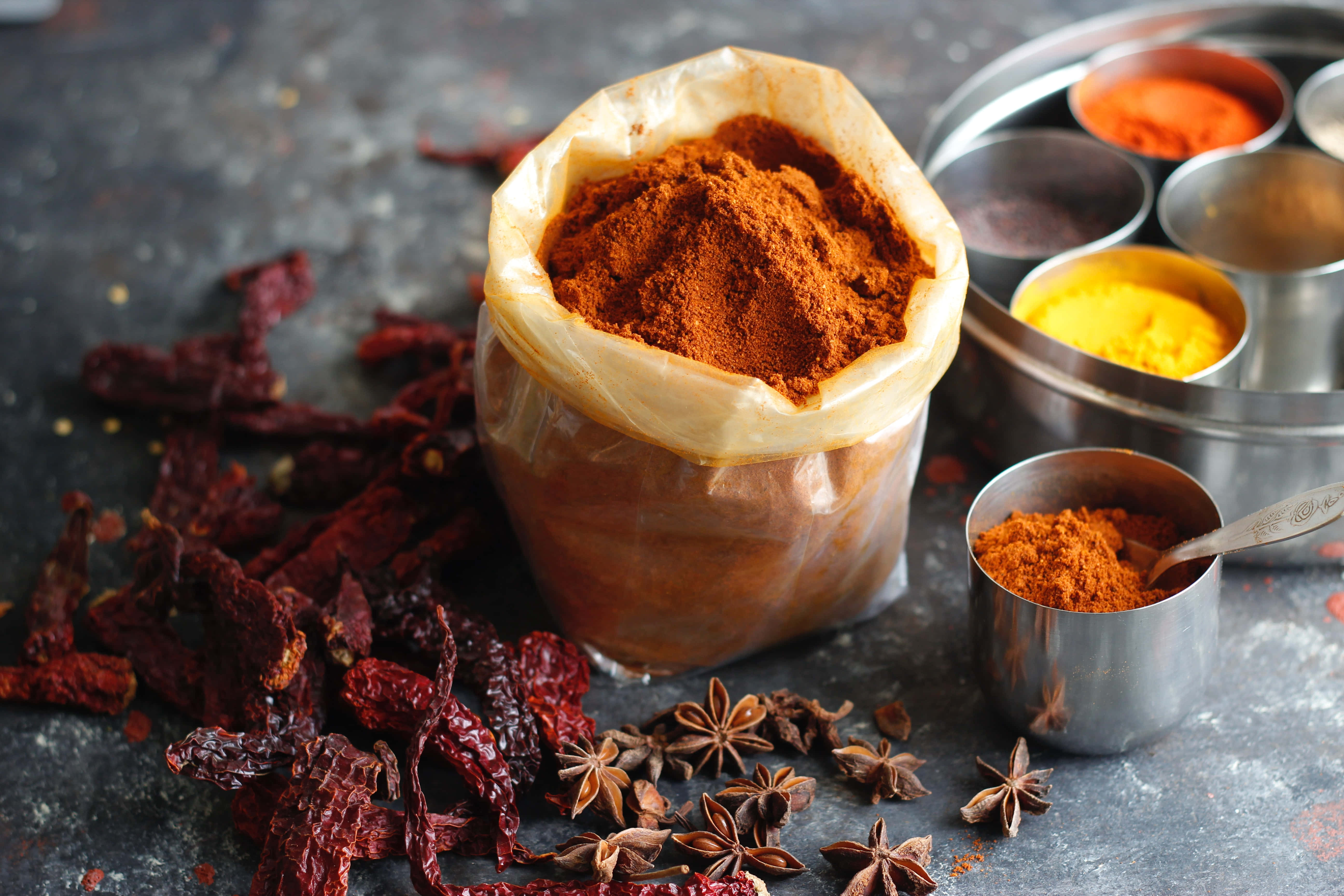 Spices 桌布 壁紙 — 一個鄉村風格的香料場景，一個紙袋裝著紅色粉末、乾辣椒和八角，散落在深色紋理表面上。