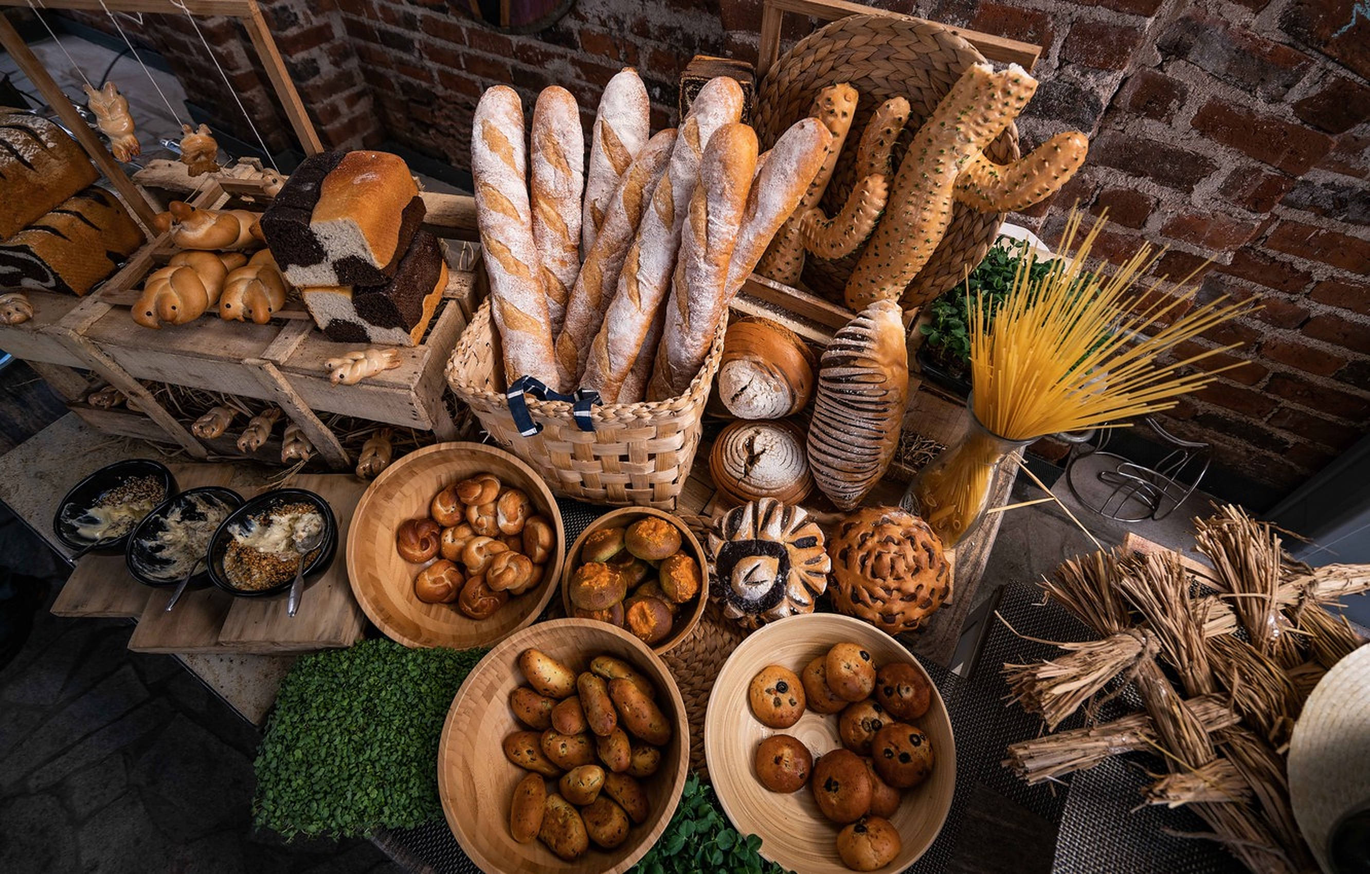 Bakery 桌布 壁紙 — 一個鄉村風格的麵包店展示，木碗裡盛著金黃色的糕點，籃子裡裝著法式長棍麵包，背景是紅磚牆。