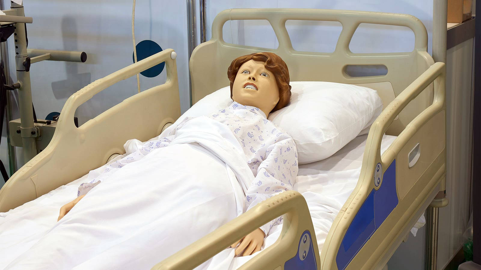 Hospital 桌布 壁紙 — 一個醫療訓練人體模型躺在醫院的床上，床上有白色床單和圖案長袍，在明亮的臨床燈光下。