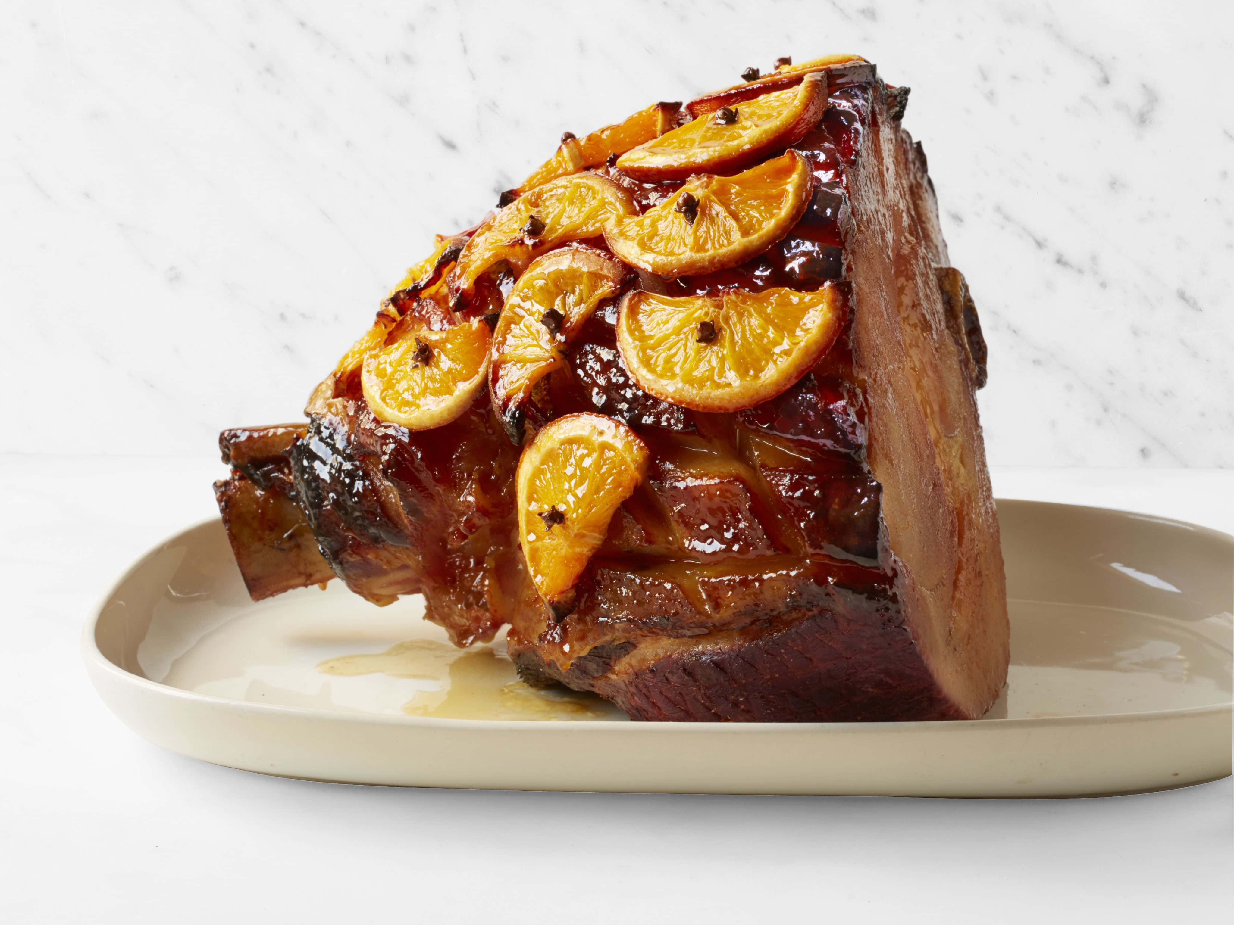 Black Forest Ham 桌布 壁紙 — 一個釉面火腿，上面放有蜜餞橙片和整粒丁香，放在米色盤子上，背景是白色大理石。