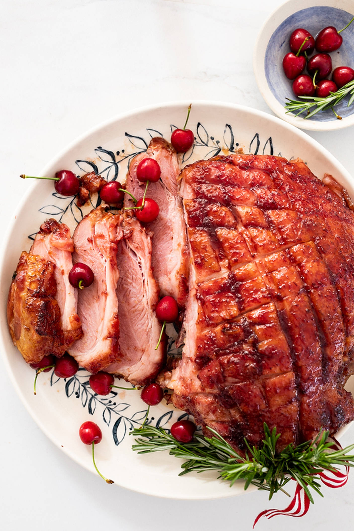 Ham 桌布 壁紙 — 一個釉面烤火腿切片放在有圖案的盤子上，用櫻桃和迷迭香裝飾，置於白色大理石表面。