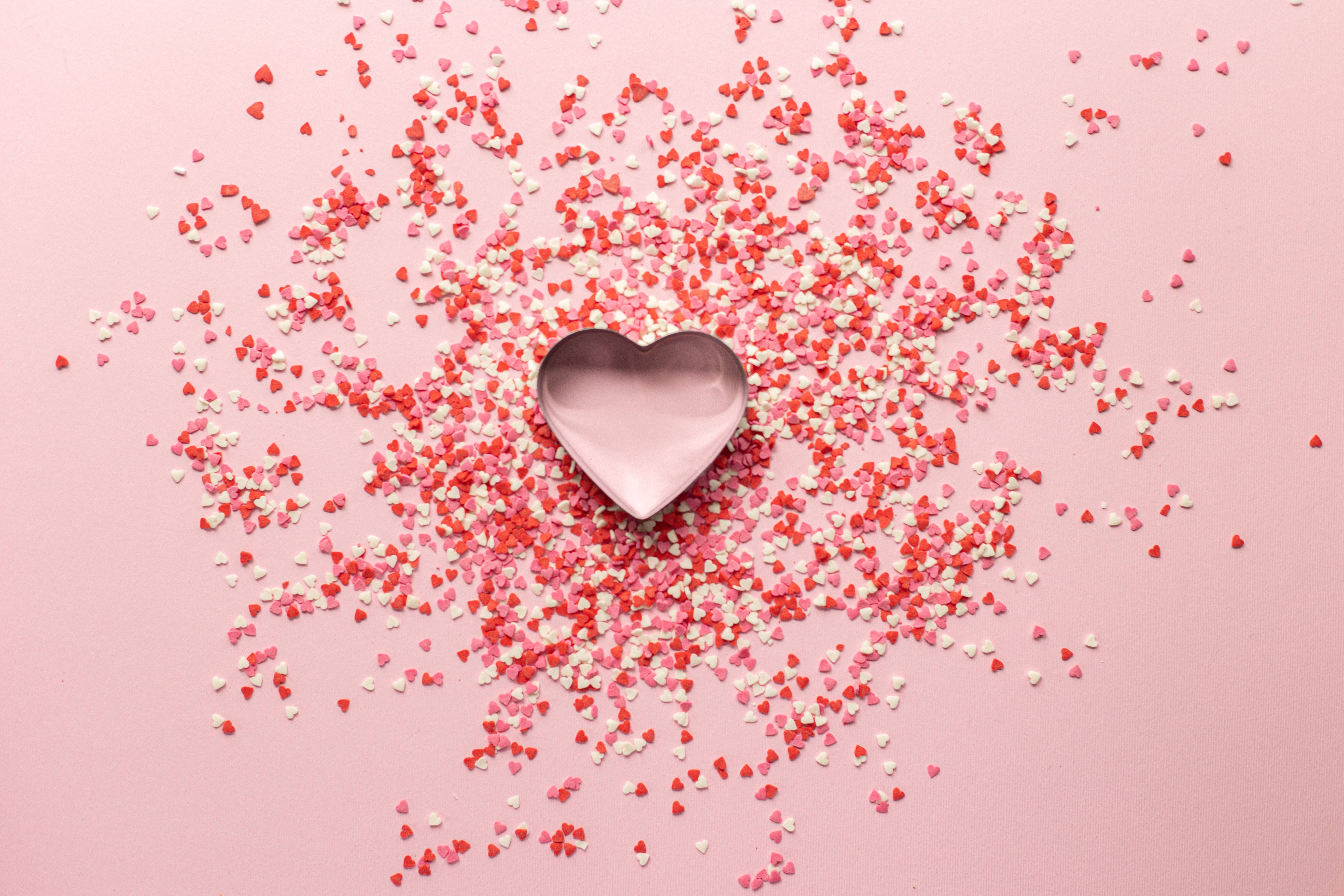 Pastel Pink Heart 桌布 壁紙 — 一個金屬心形餅乾模具，周圍散落著紅色和白色的心形糖粒，背景是柔和的粉紅色。