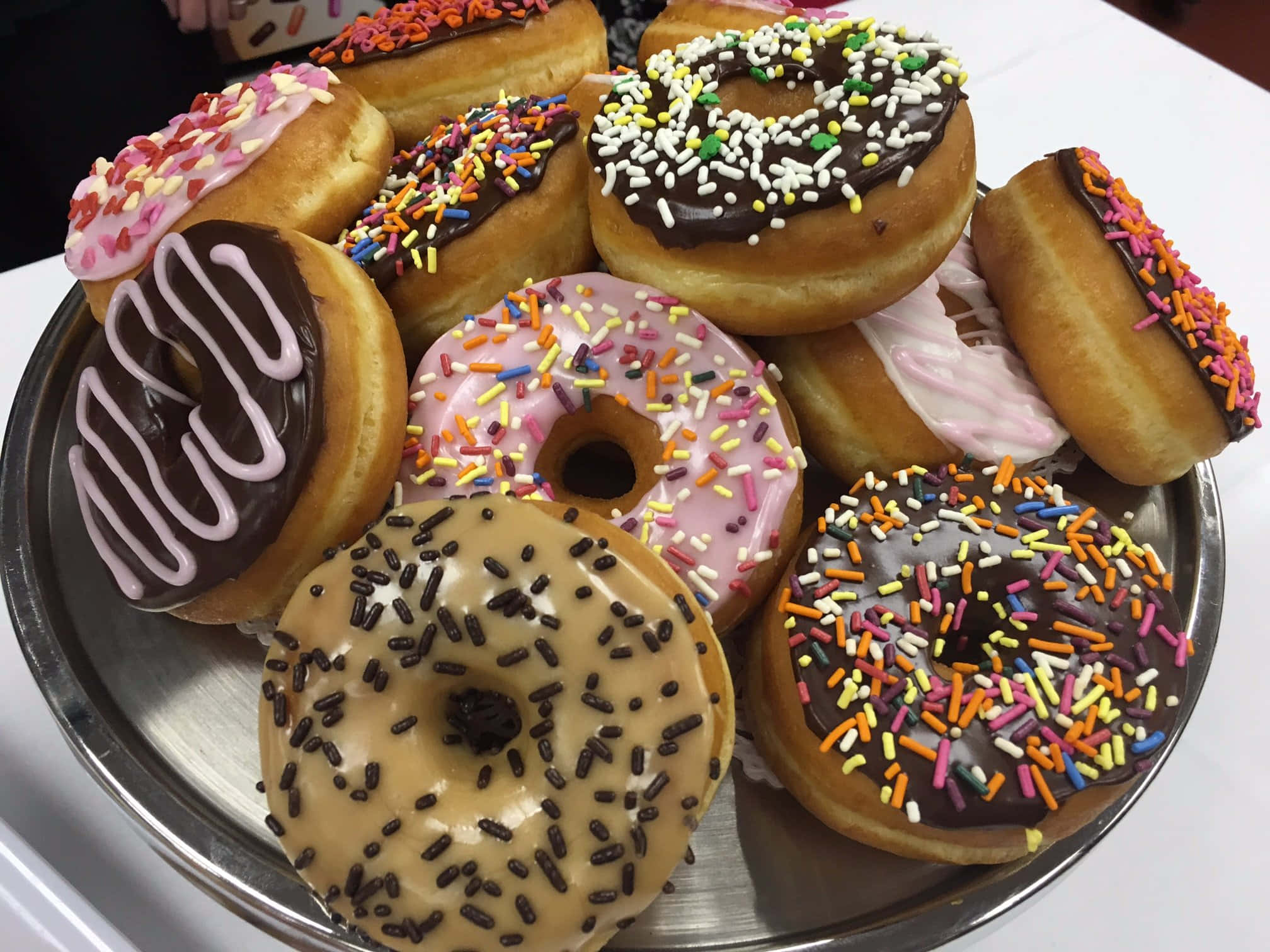 Dunkin Donuts 桌布 壁紙 — 一個金屬托盤上堆滿了各式各樣的糖霜甜甜圈，上面撒滿了彩色糖粒和巧克力醬。