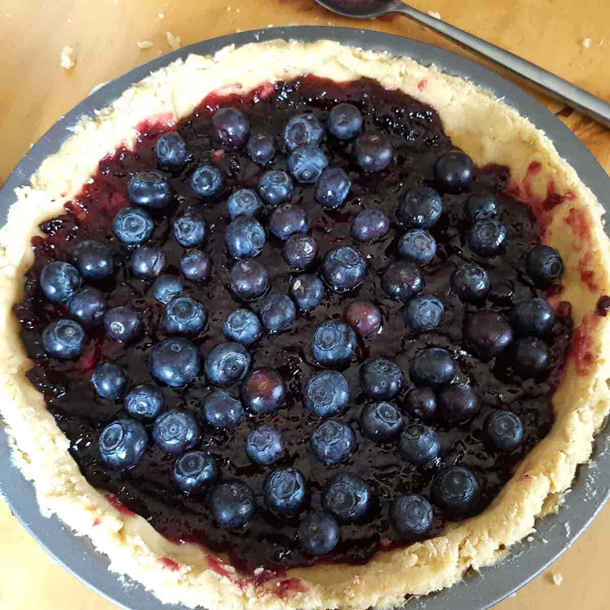 Blueberries Tart 桌布 壁紙 — 一個金屬盤中的藍莓派特寫，上面有新鮮藍莓和深色糖漿，放在木桌上。