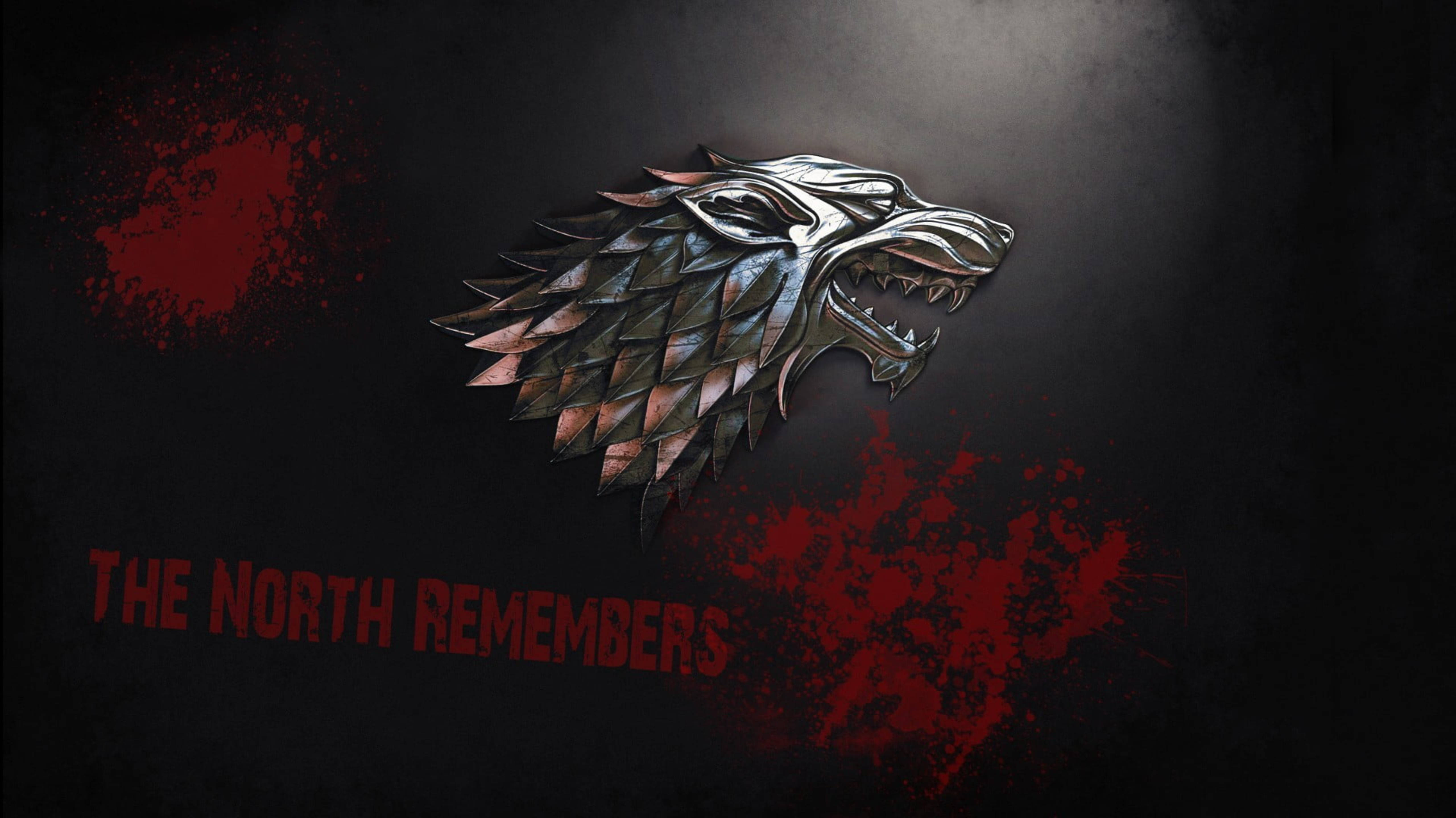 House Stark 桌布 壁紙 — 一個金屬製的冰原狼頭徽章，帶有紅色飛濺物和「THE NORTH REMEMBERS」字樣，背景為深色。
