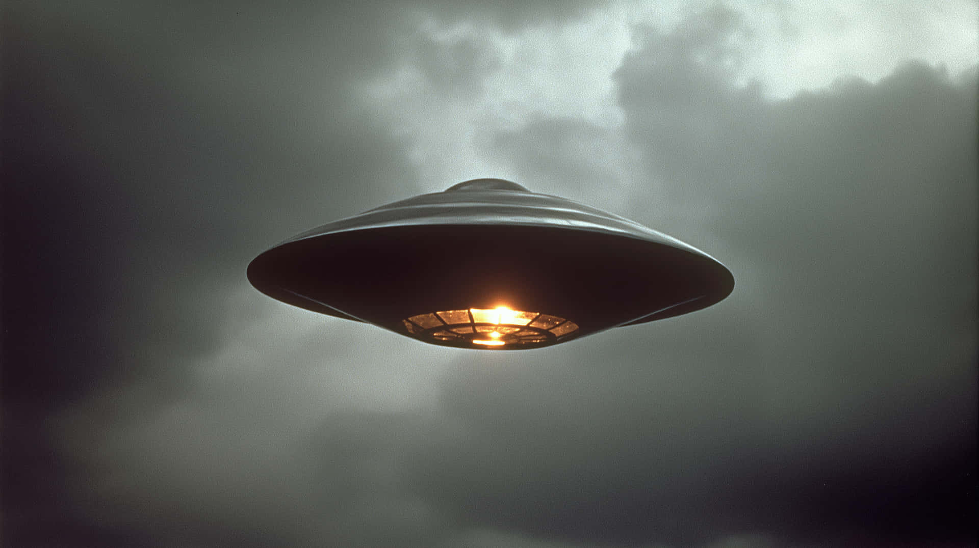 Glowing Object in Overcast Sky, Mysterious Encounter 壁紙 — 一個金屬飛碟在暴風雨般的灰色天空下盤旋，中心發出脈動的橙色光芒。