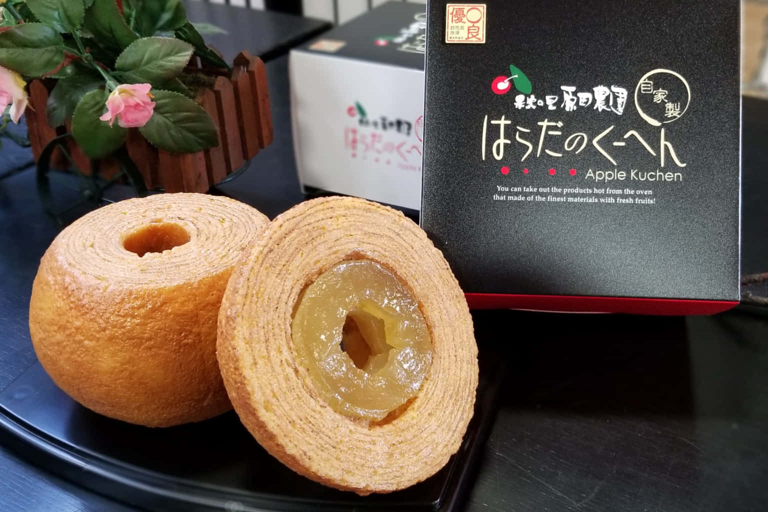 Japanese Sweets 桌布 壁紙 — 一個金棕色蘋果庫亨糕點切開，露出閃亮的蘋果餡料，旁邊放著黑色包裝和一朵粉紅色的花。