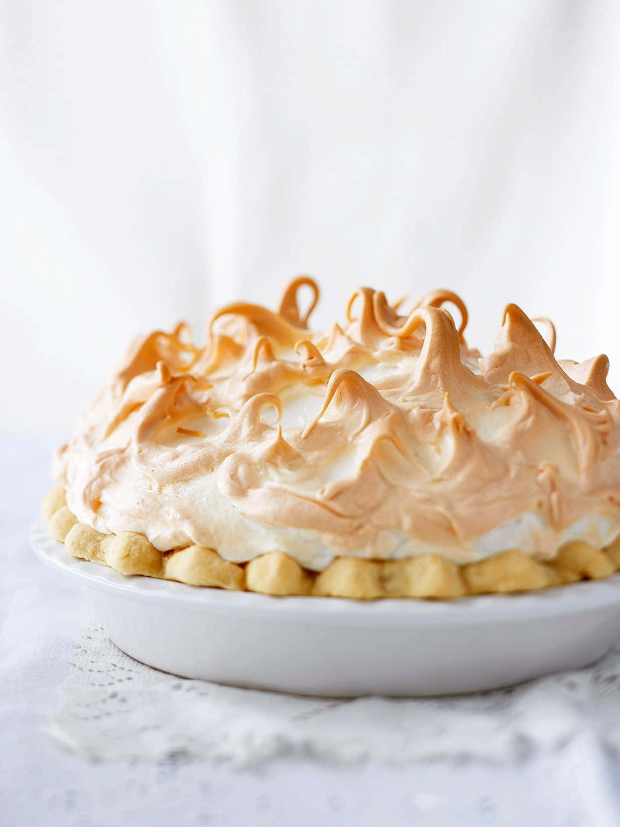 Meringue 桌布 壁紙 — 一個金棕色蛋白霜派，帶有凹槽邊緣，放在白色盤子上，盤下墊著蕾絲餐墊，背景是柔和的白色。