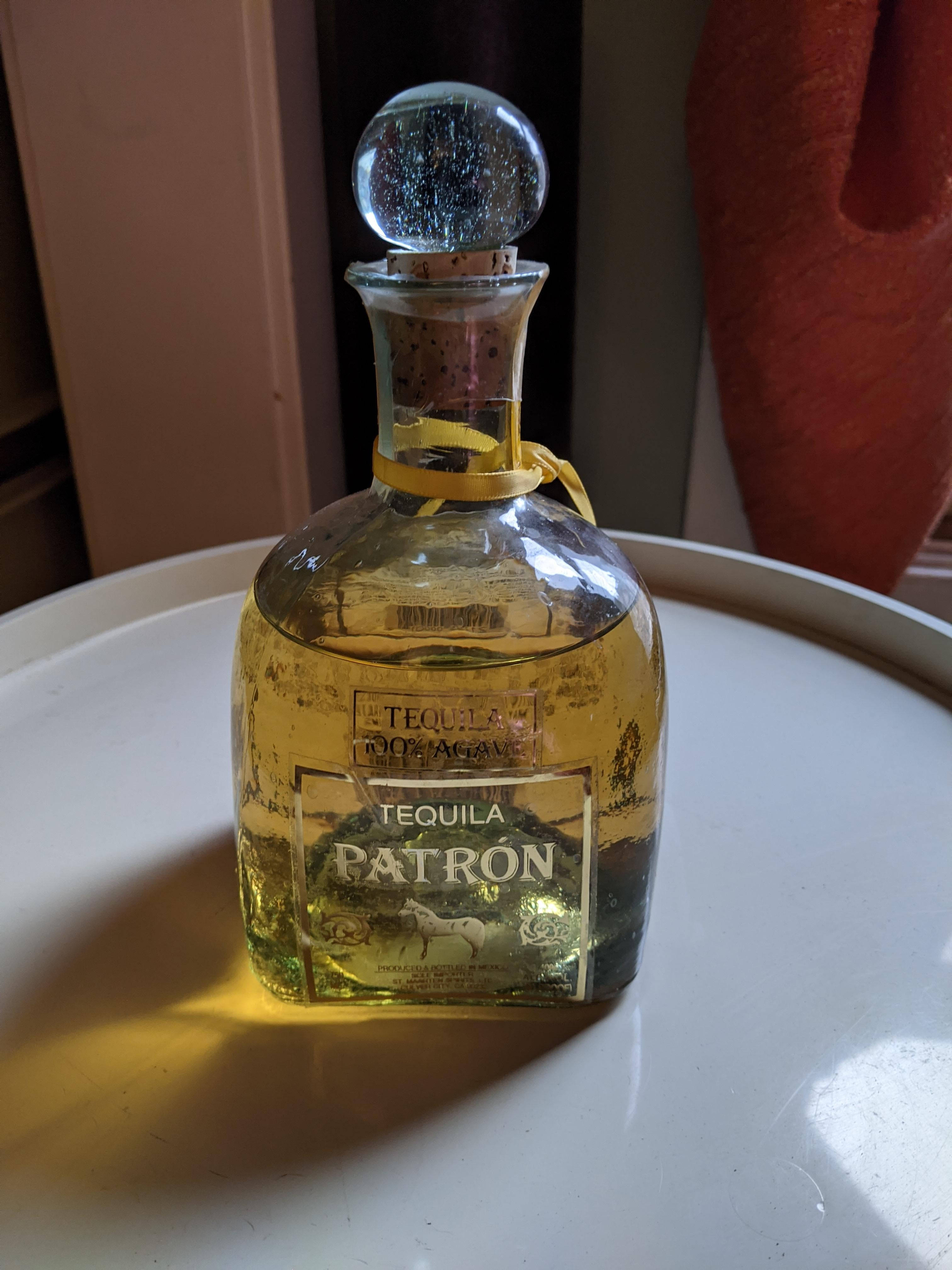 安卓 桌布 壁紙 — 一個金色Patron龍舌蘭酒瓶，帶有水晶塞，放在一個沐浴在溫暖陽光下的白色托盤上。