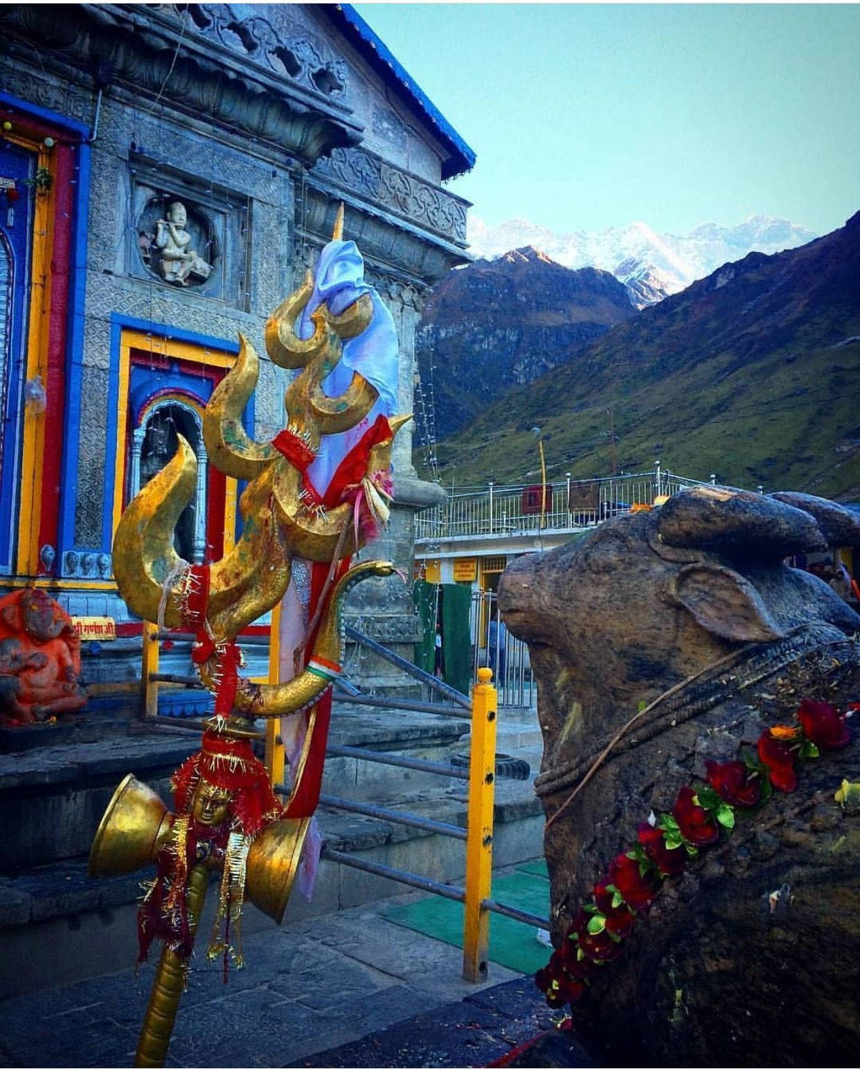 Indian 桌布 壁紙 — 一個金色三叉戟裝飾著紅色絲帶，矗立在色彩繽紛的寺廟前，背景是積雪的山脈。