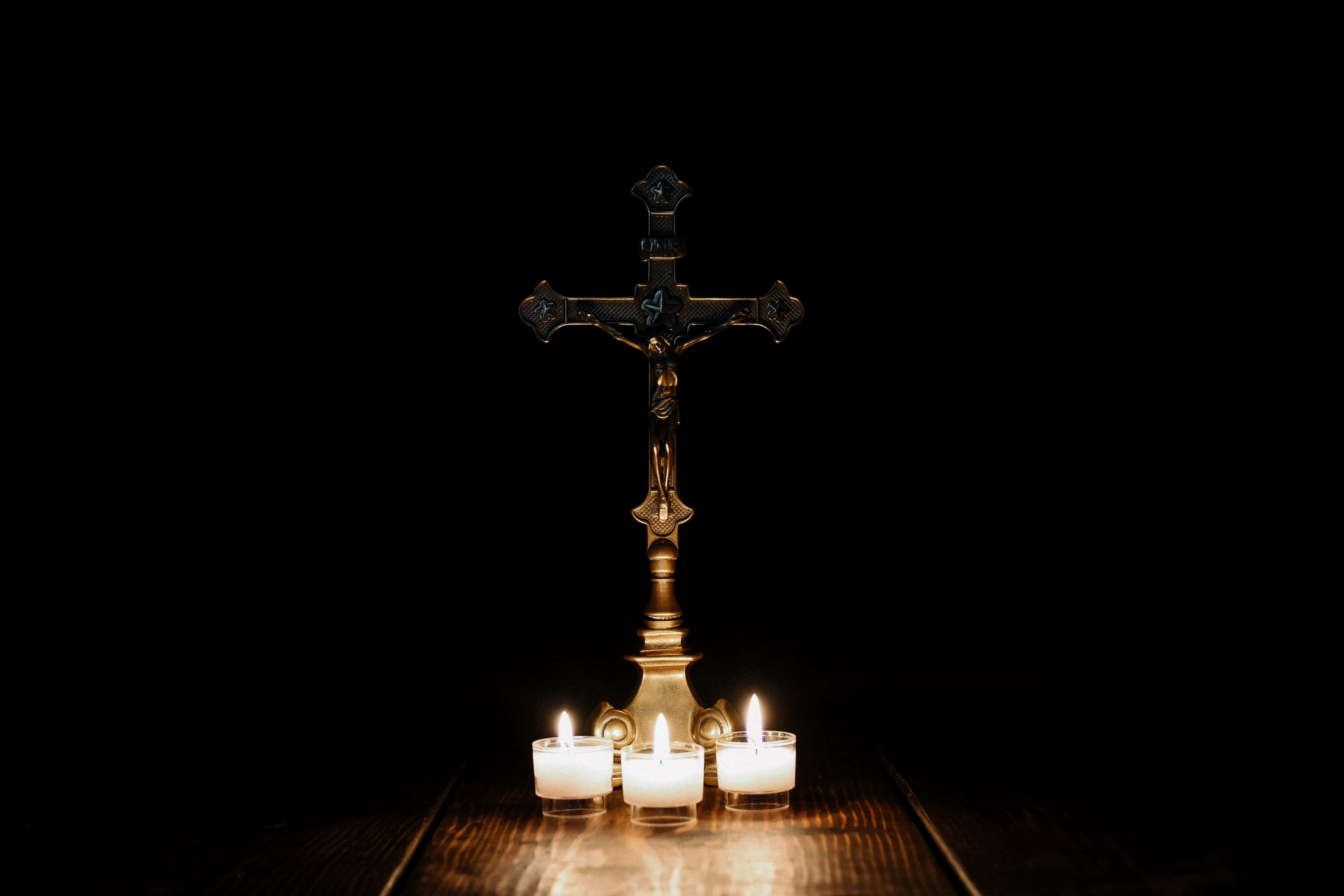 Catholic 桌布 壁紙 — 一個金色十字架莊嚴地立在木質表面上，由三根搖曳的蠟燭照亮，背景漆黑一片。