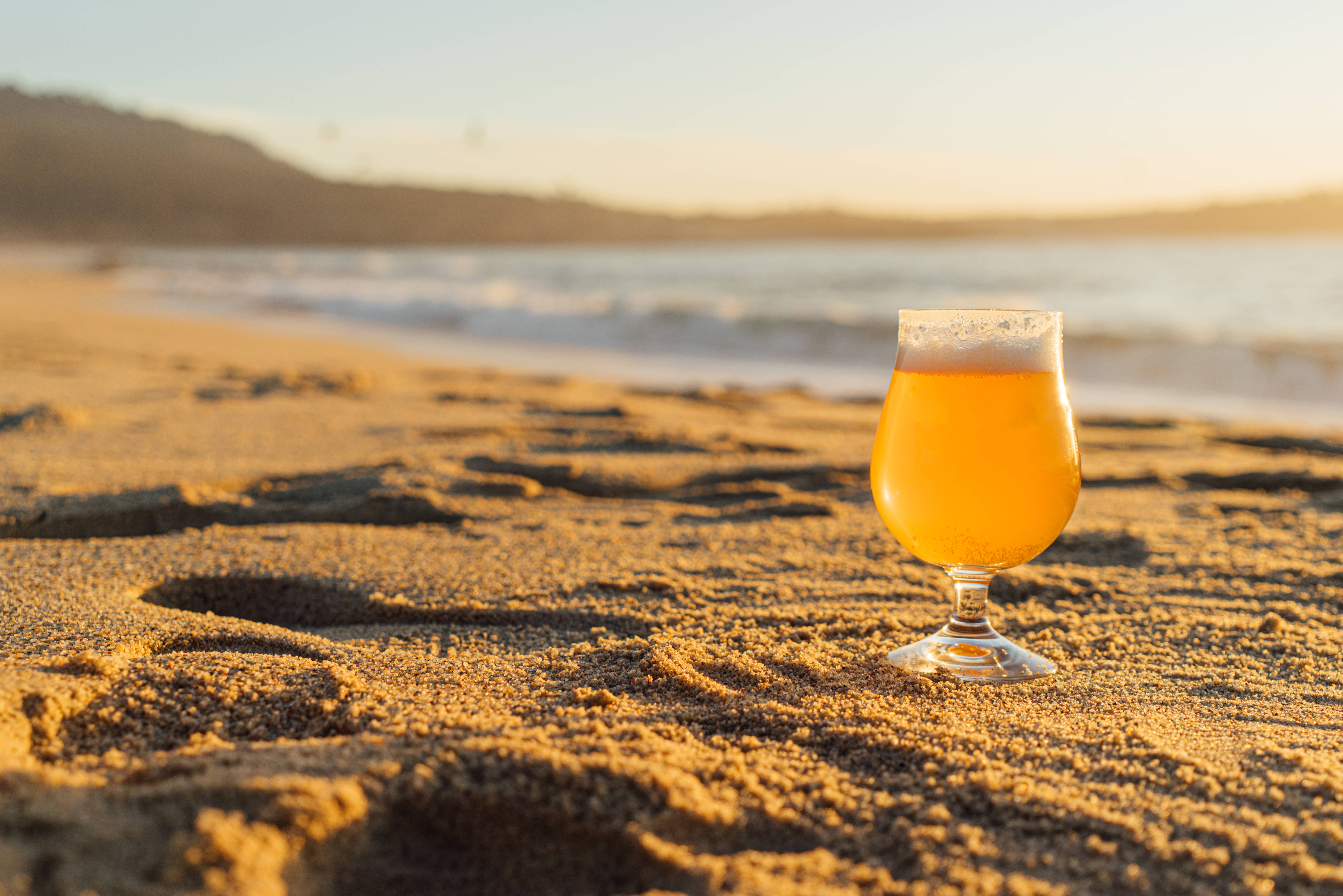 Beer 桌布 壁紙 — 一個金色啤酒杯放在溫暖的沙灘上，夕陽下背後有輕柔的海浪湧來。