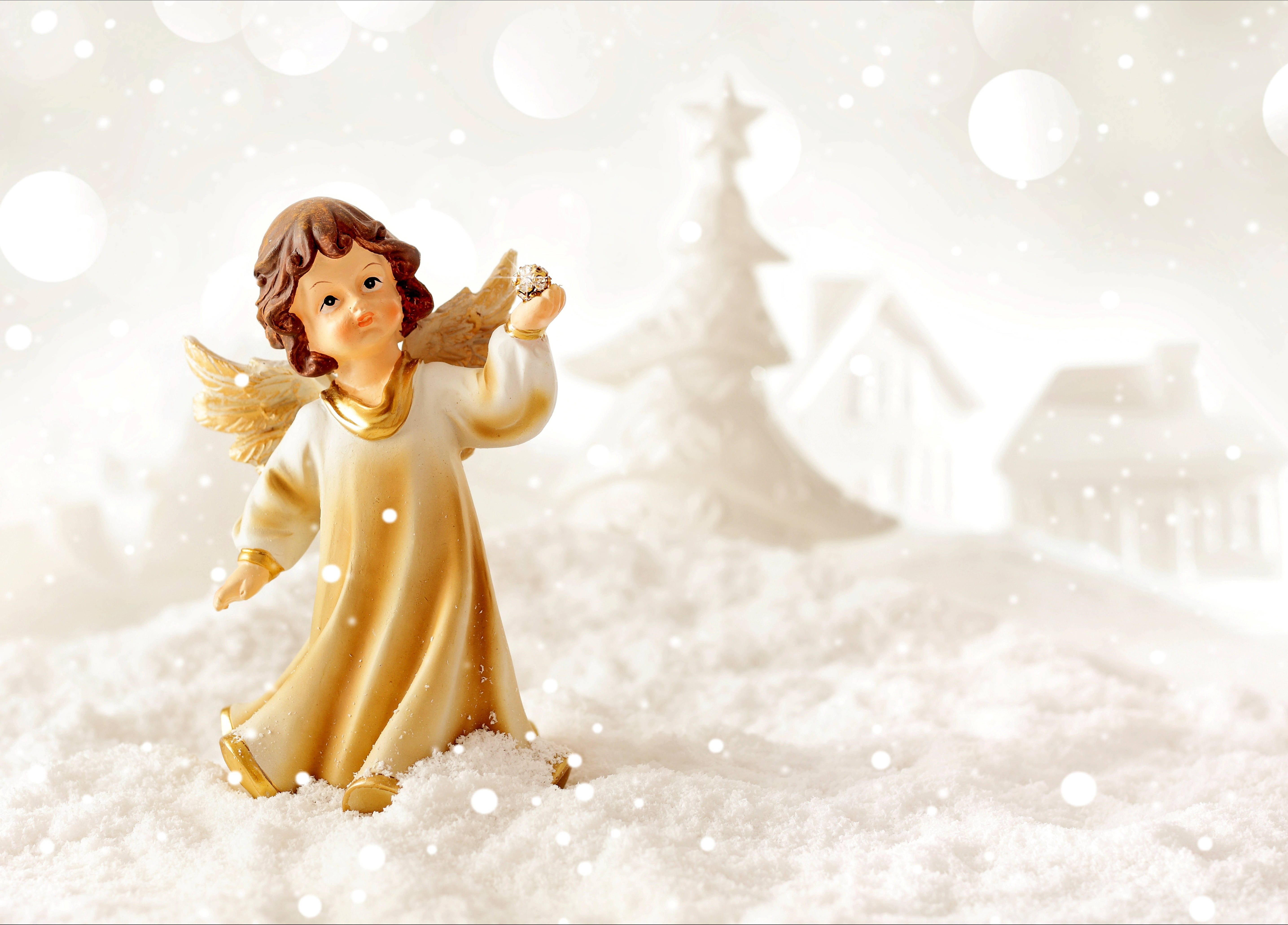 angels 桌布 壁紙 — 一個金色天使雕像站在雪景中，背景有模糊的房屋和樹木，在柔和的光芒下。