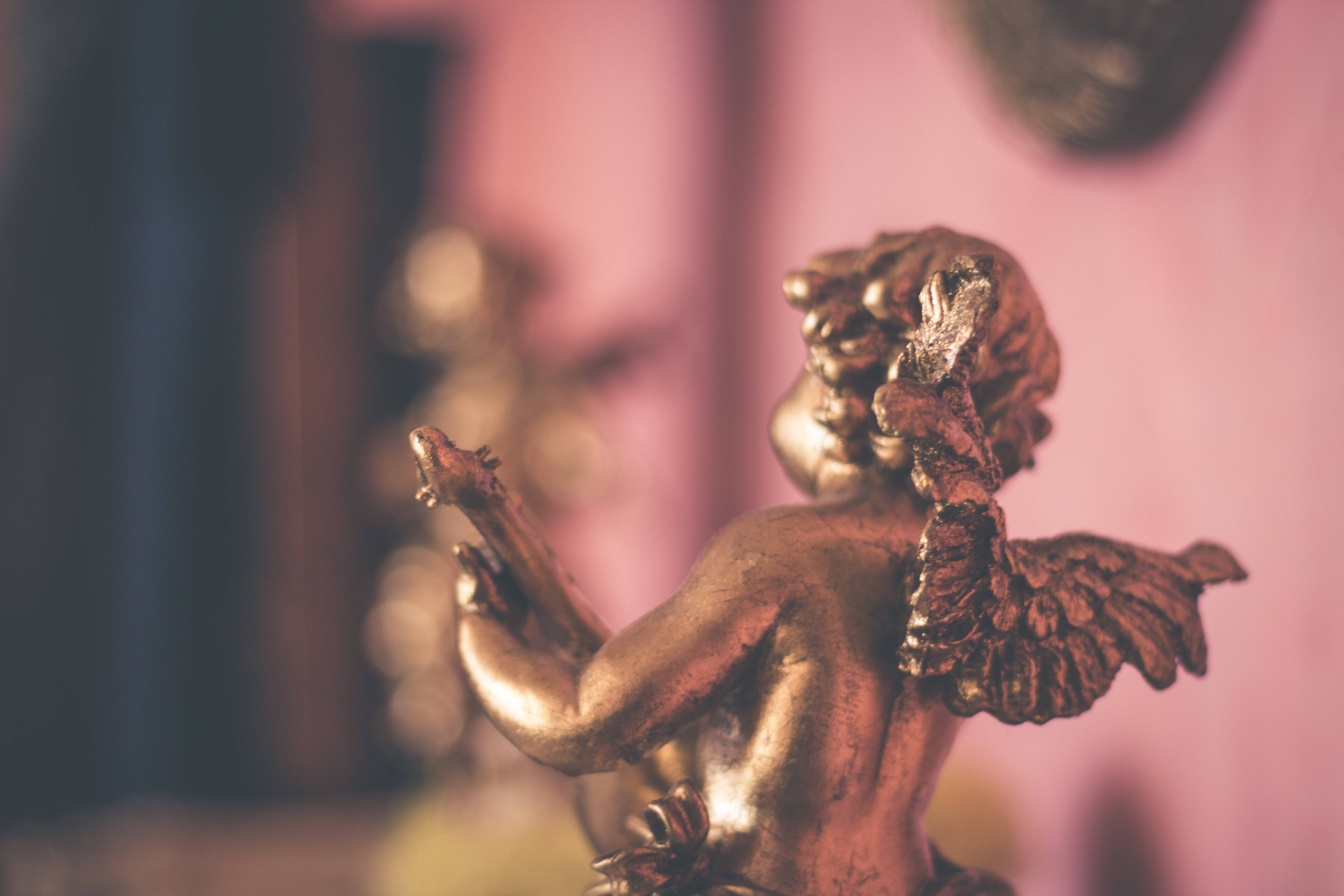 angels 桌布 壁紙 — 一個金色小天使雕像，帶有精細的翅膀和俏皮的姿勢，站在柔和的粉紅色光線中，背景模糊。