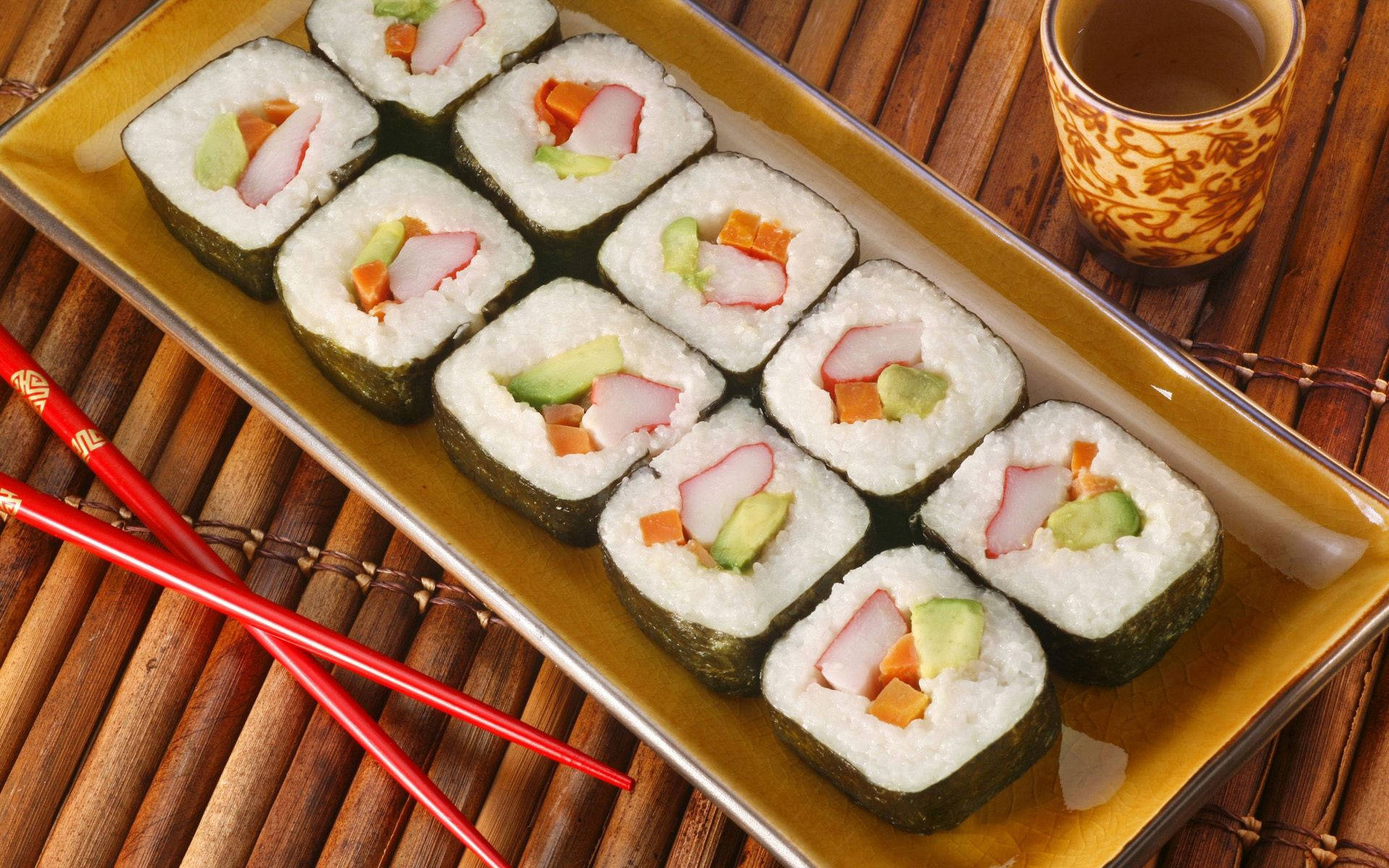 Sushi 桌布 壁紙 — 一個金色托盤上整齊排列著八個壽司卷，內有粉紅色魚肉、酪梨和紅蘿蔔，放在竹蓆上，旁邊有紅色筷子及有圖案的茶杯。