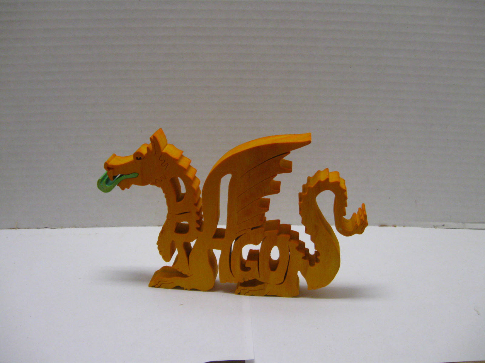 Gold Dragon 桌布 壁紙 — 一個金色木龍公仔，有綠色舌頭，站在白色表面上，背景是灰色波浪紋。