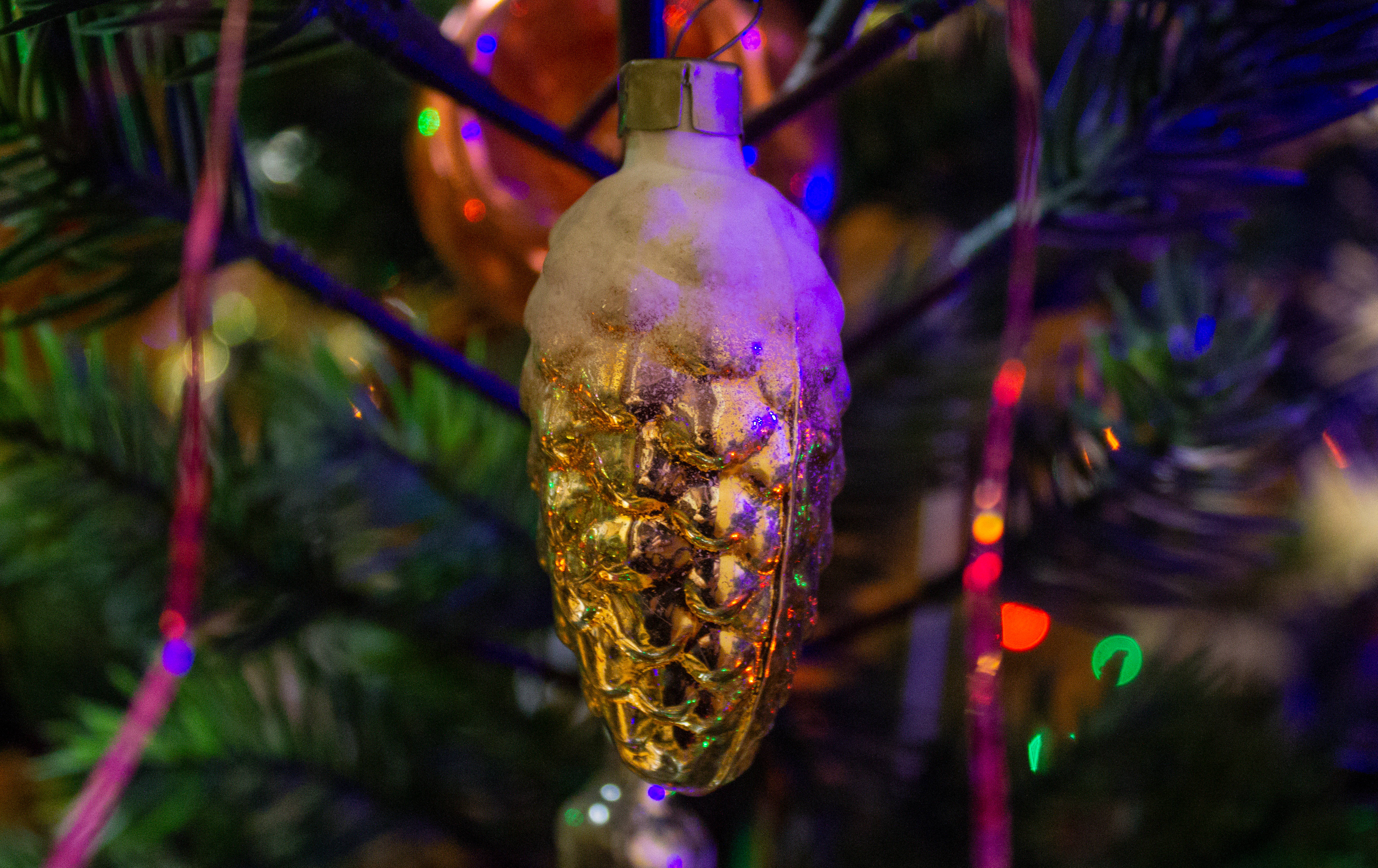 聖誕美學 桌布 壁紙 — 一個金色松果形狀的裝飾品，上面灑滿了雪，掛在掛滿彩色燈飾的聖誕樹上的特寫。