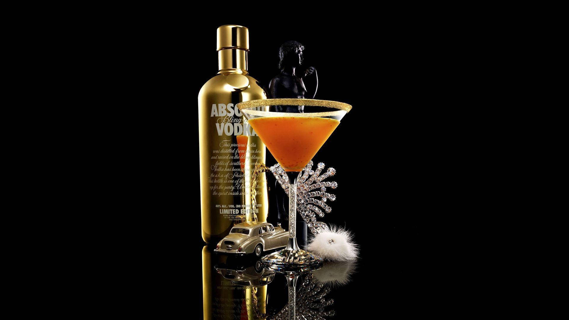 bottle 桌布 壁紙 — 一個金色的Absolut Vodka酒瓶旁邊放著一杯充滿活力的橙色雞尾酒，盛在飾有水晶和 fluffy 白色絨球裝飾的馬提尼杯中，置於黑色反光表面上。