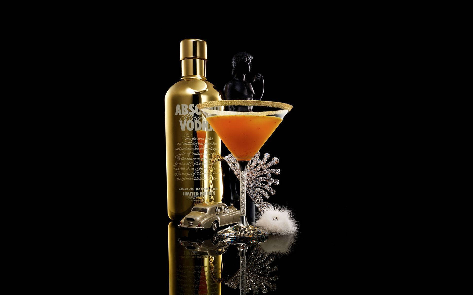 飲料 桌布 壁紙 — 一個金色的Absolut Vodka酒瓶放在一個裝滿橙色雞尾酒的馬丁尼酒杯旁邊，酒杯上飾有水晶和羽毛，置於黑色反光表面上。
