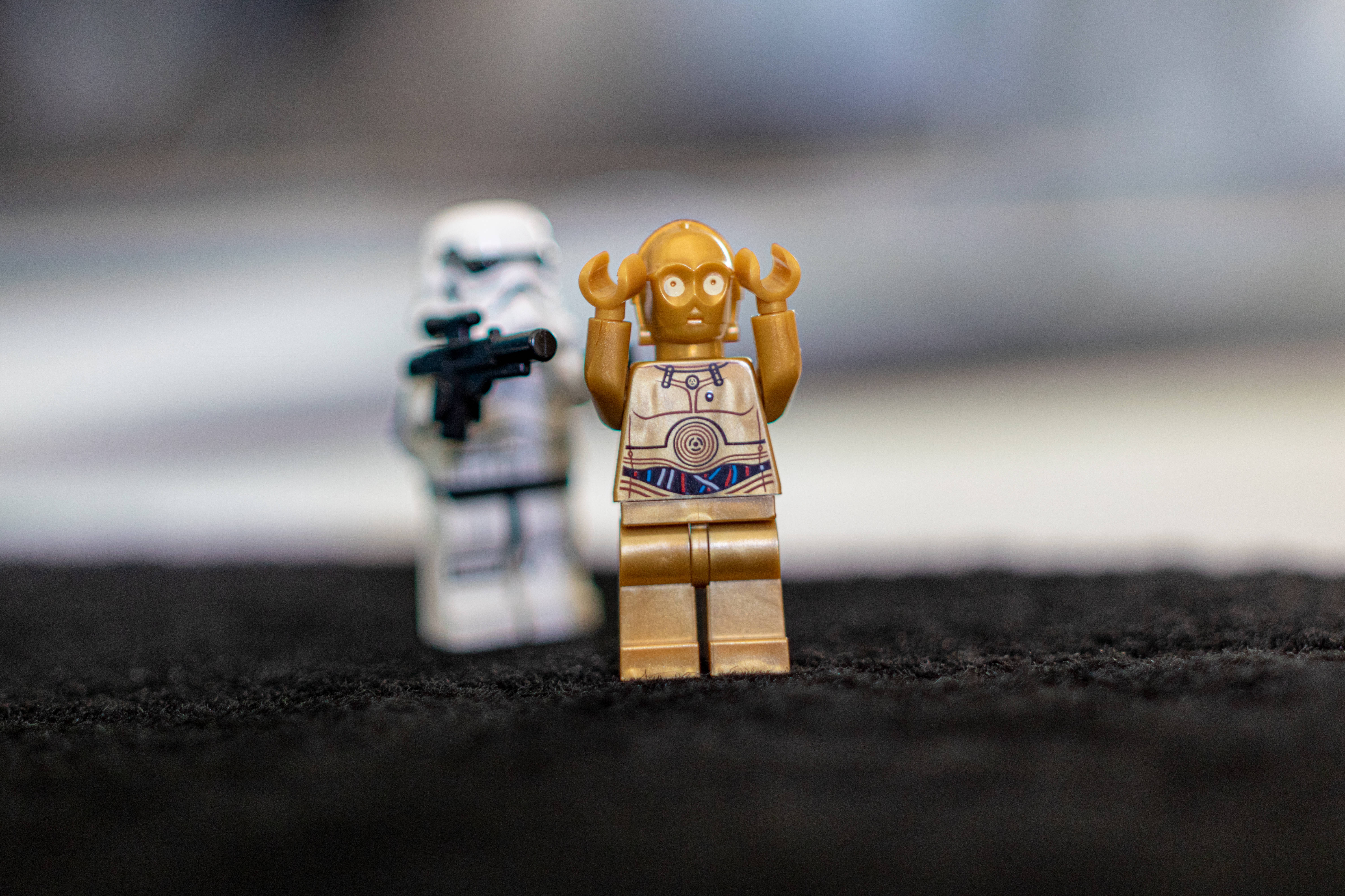 暴風兵 桌布 壁紙 — 一個金色的C-3PO LEGO迷你人偶站在黑色紋理表面上，表情驚訝，後面有一個模糊的風暴兵。