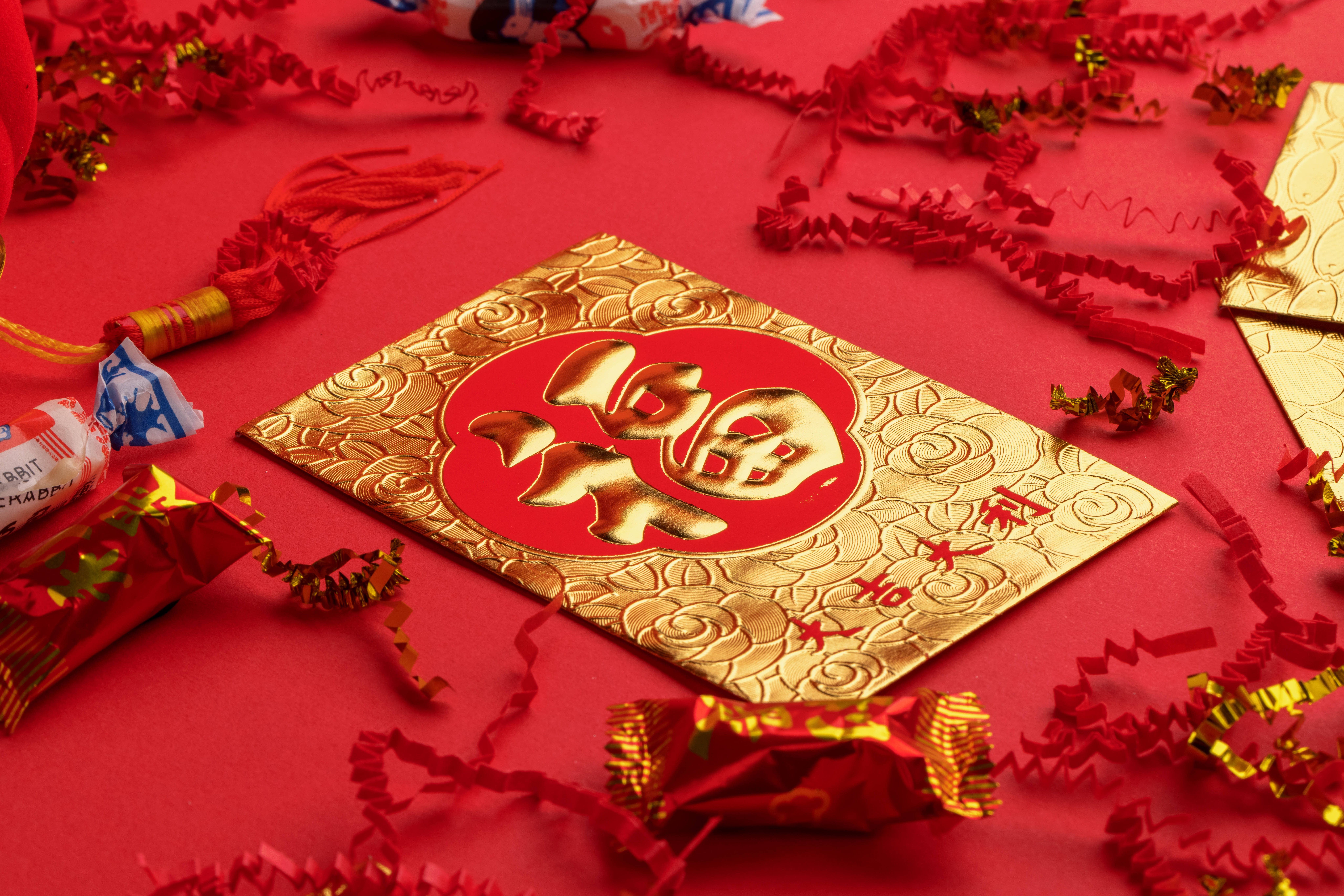 春節 桌布 壁紙 — 一個金色的中式紅包，上面有浮雕的「福」字，放在鮮紅色的表面上，周圍是喜慶的紅色和金色紙屑以及包裝好的糖果。