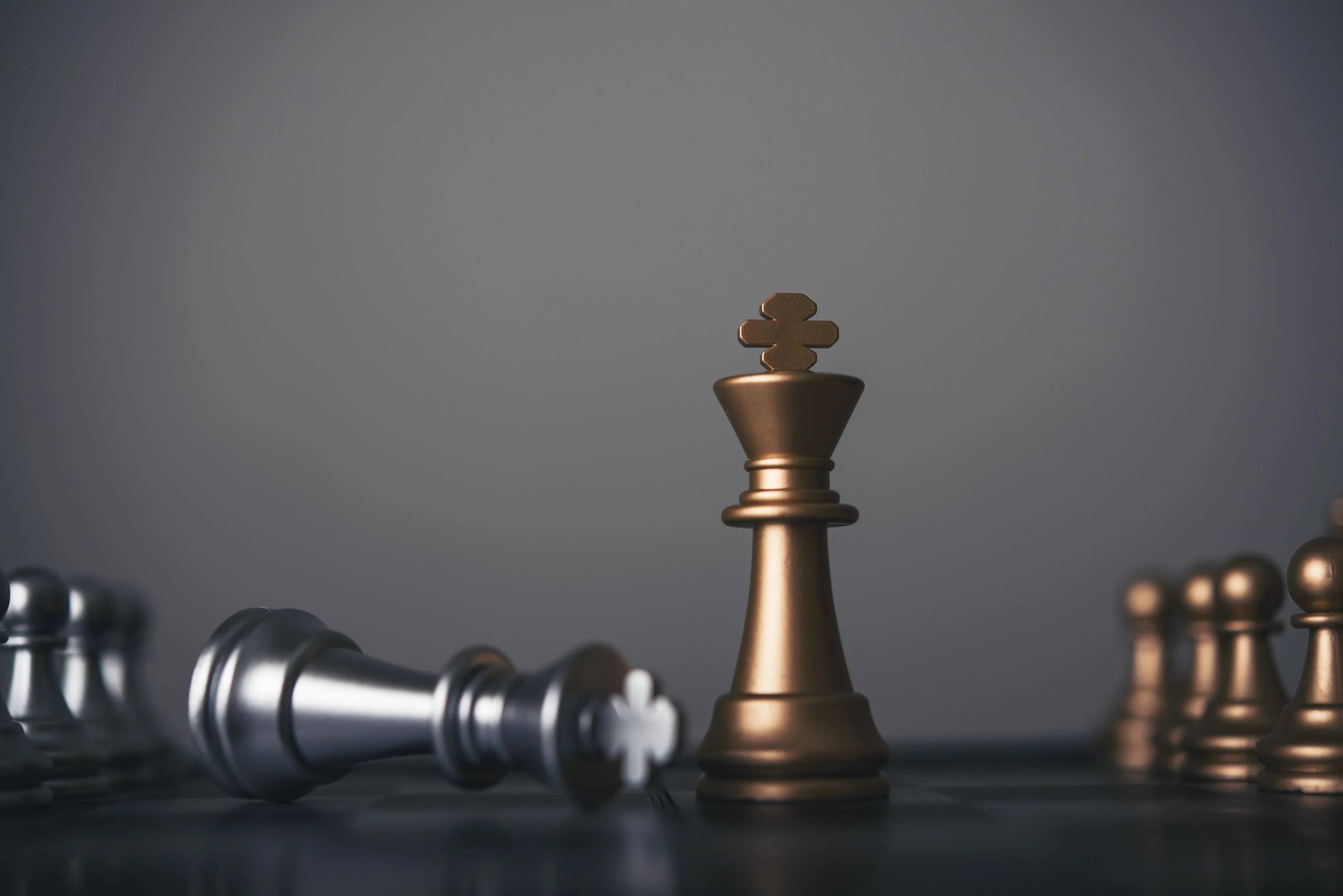 西洋棋 桌布 壁紙 — 一個金色的西洋棋國王高高站立在深色棋盤上，在柔和的灰色背景前，立於倒下的銀色棋子之上，充滿勝利的姿態。