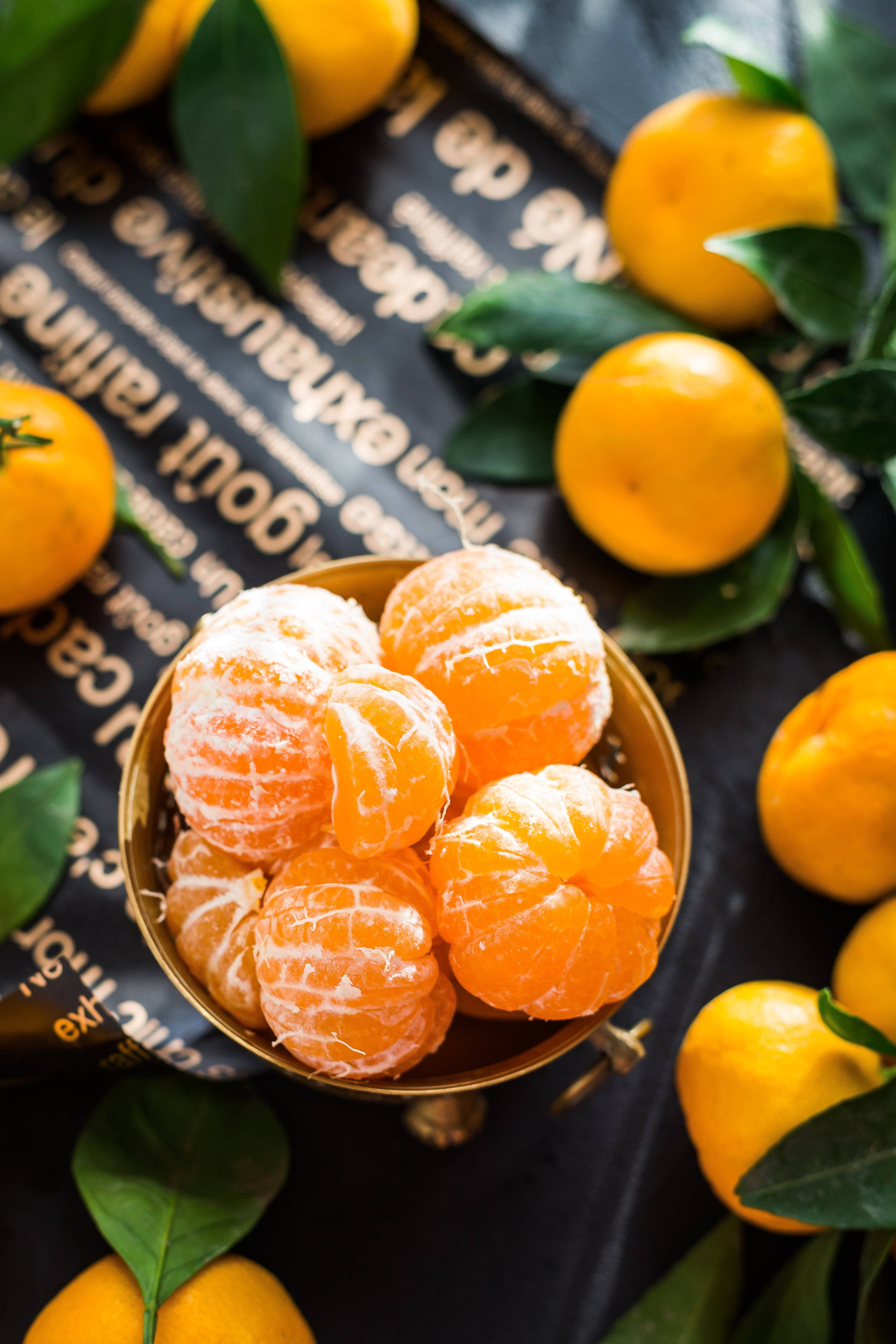 Food Photography 桌布 壁紙 — 一個金色碗裝滿剝皮柑橘，周圍有完整水果和綠葉，放在深色紋理表面上。