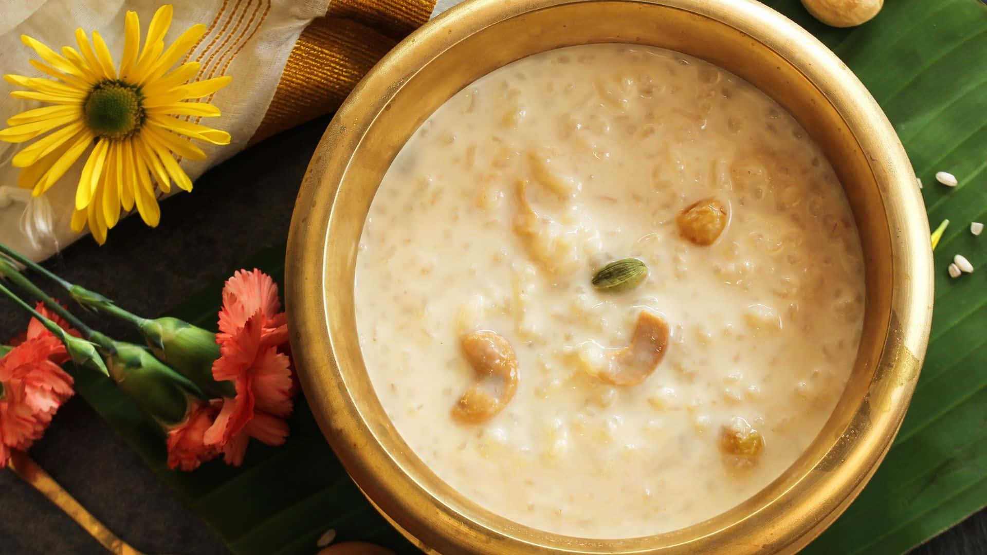 Vishu 桌布 壁紙 — 一個金色碗裝著奶油狀的kheer，上面放有腰果和荳蔻，放在香蕉葉上，旁邊有鮮黃色和粉紅色的花朵。