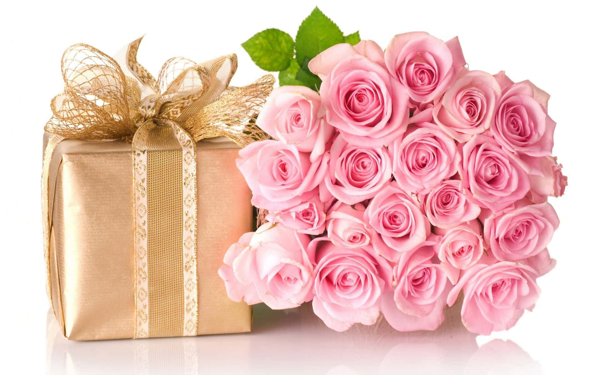 Birthday Message 桌布 壁紙 — 一個金色禮物盒，帶有閃閃發光的蝴蝶結，旁邊是一束柔和的粉紅玫瑰，配有綠葉，放在反光的白色表面上。